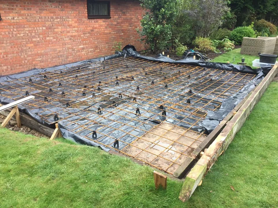 Construction site with rebar grid installed on a black plastic sheet, surrounded by wooden formwork, next to a brick house and green lawn.