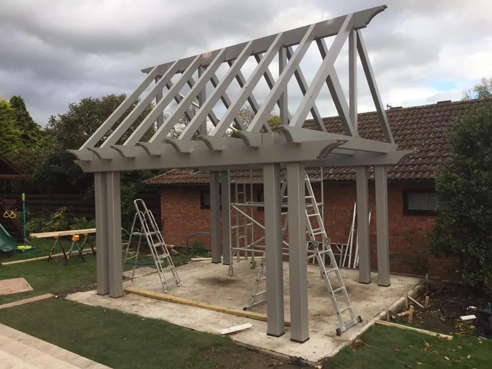 Construction of a wooden pergola with vertical posts and roof beams in a backyard, with ladders and construction tools around.