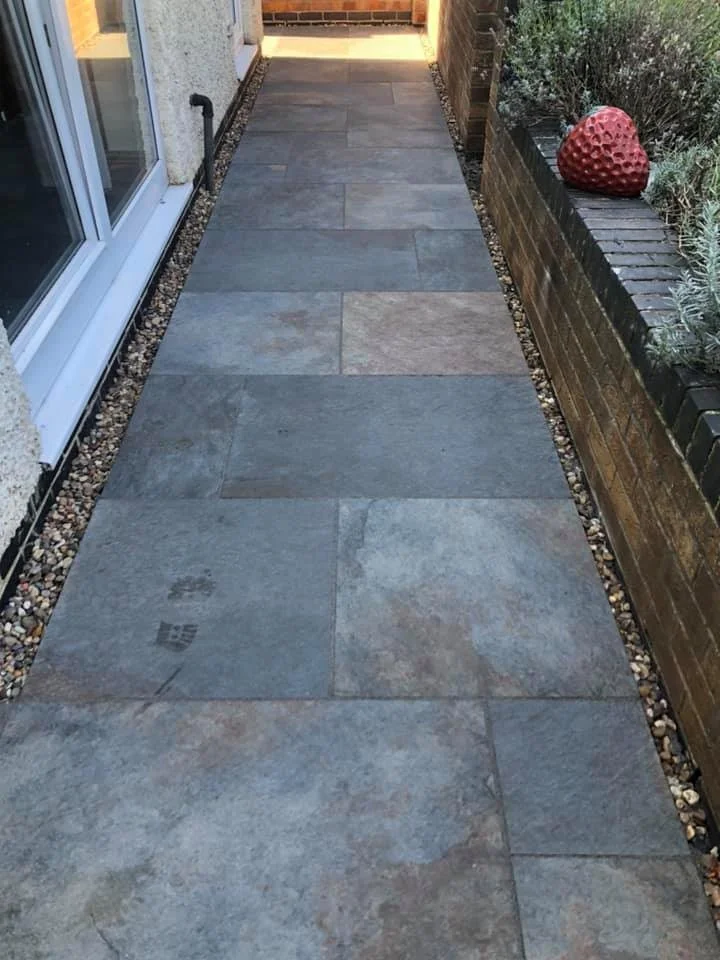A narrow outdoor pathway made of large rectangular gray and beige stone tiles, bordered by small pebbles, next to a brick wall with a decorative red object and some plants.