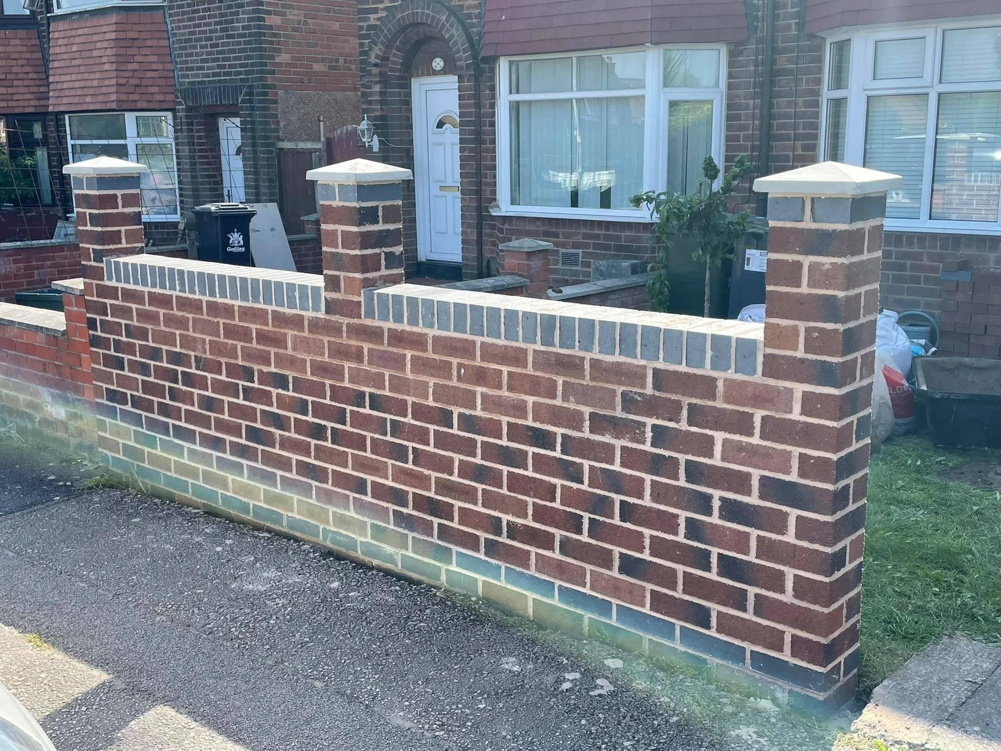 Front view of a brick house with a brick fence, white window, and white front door, with a small patch of grass and some bags and items near the house.