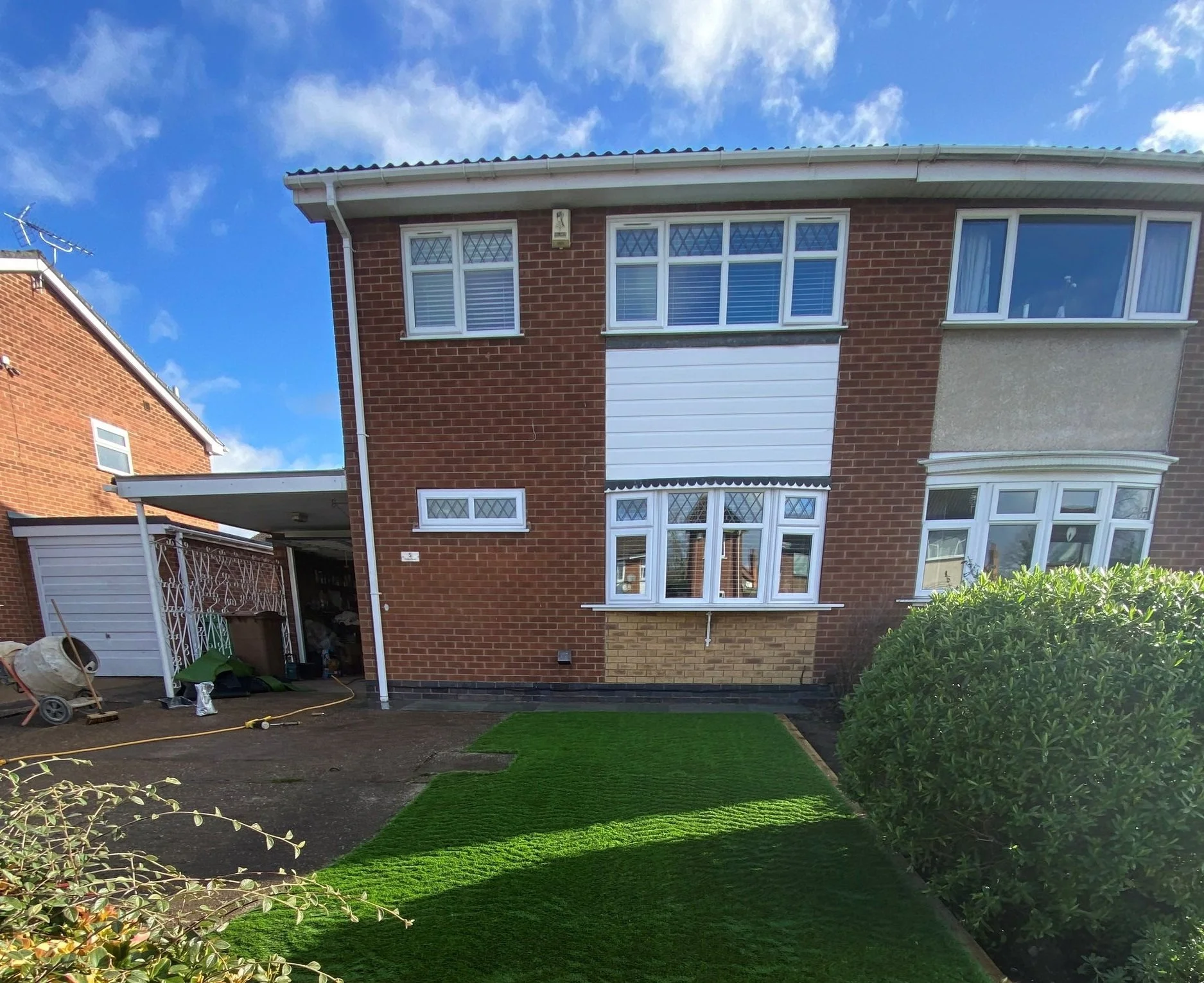 Rear view of a brick house with multiple windows, a lawn, and a adjacent garage with outdoor tools, against a partly cloudy sky.