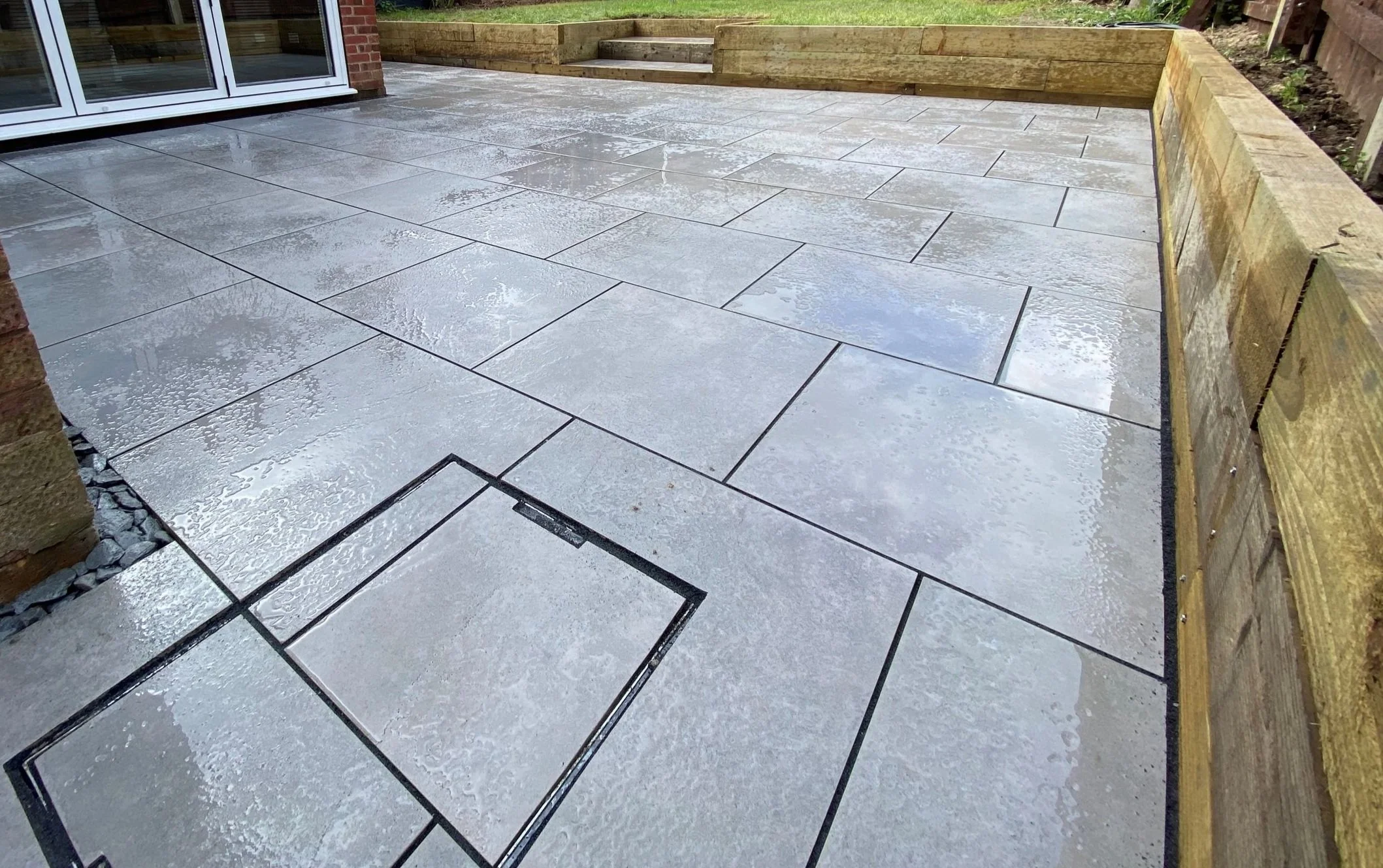 Wet grey stone patio with a wooden raised garden bed along the edge, and a sliding glass door to a house in the background.