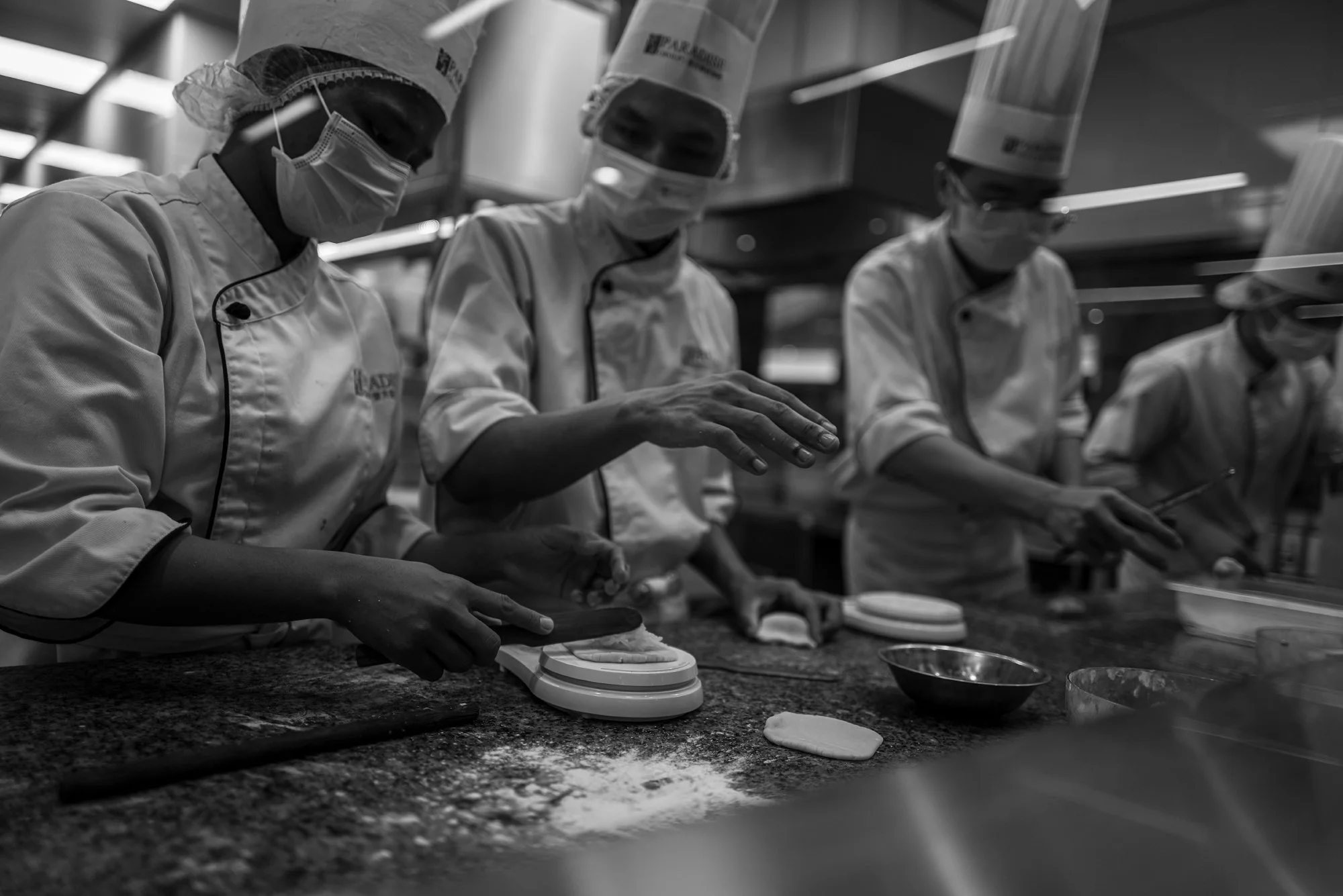 Print AD-0053. Culinary workers in masks and chef uniforms preparing dough on a commercial kitchen counter.