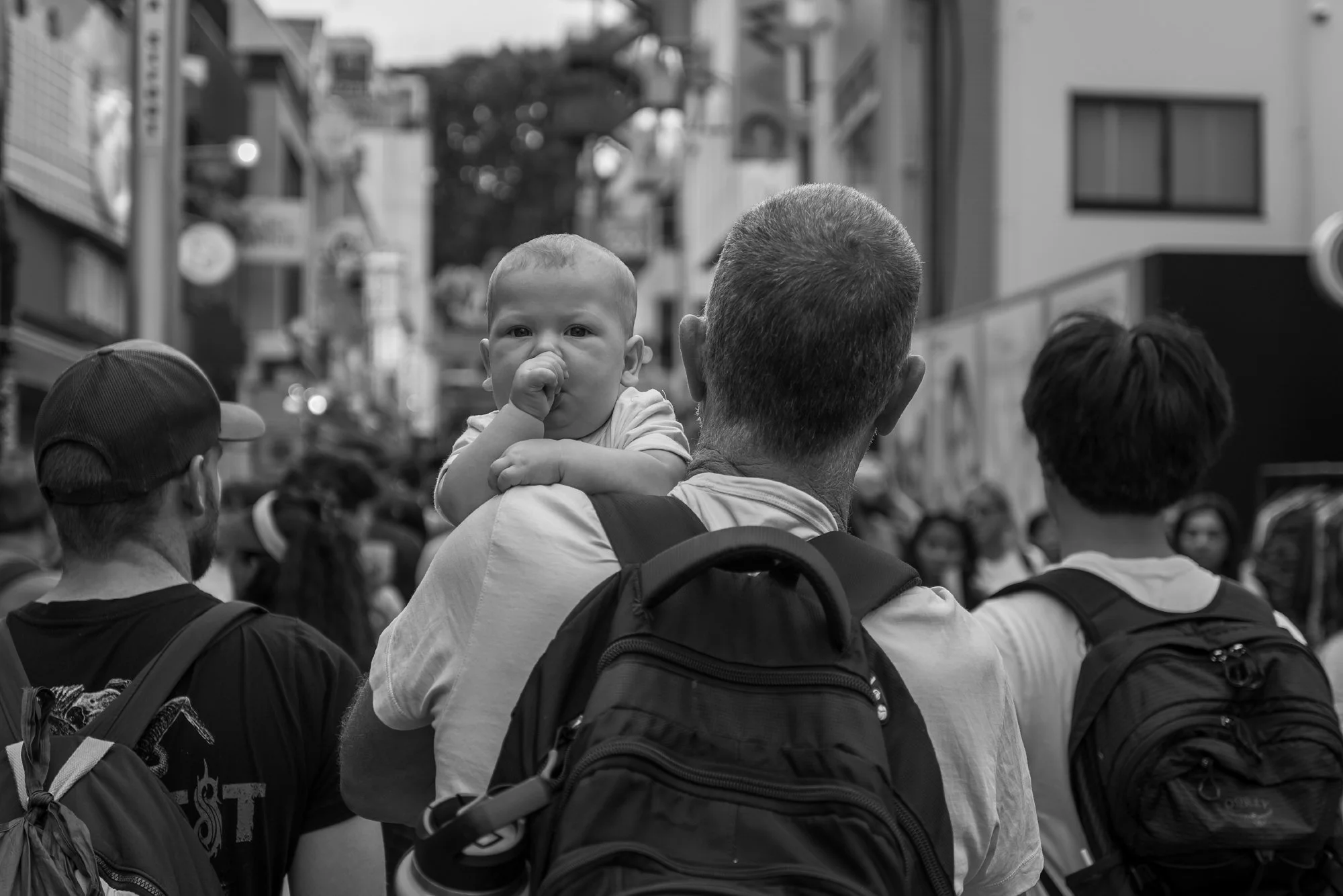 Print AD-0031. A baby looking back over an adult’s shoulder in a crowd.