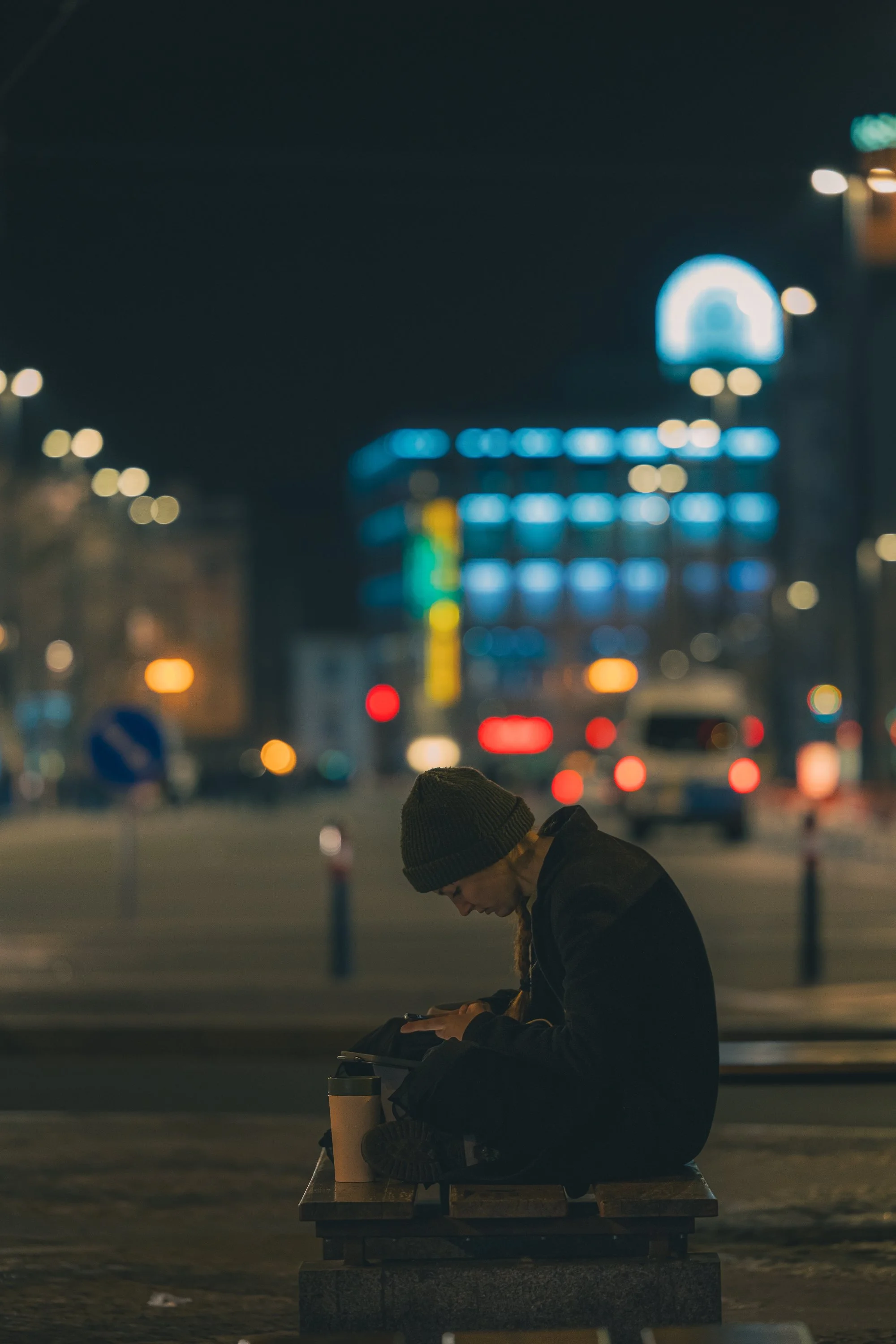 Print AD-0047. A person seated on a bench at night against blurred city lights.