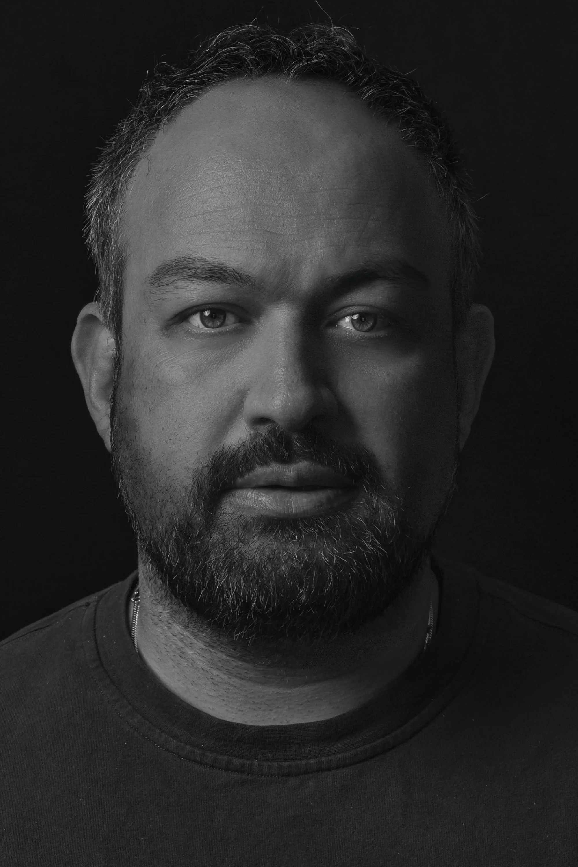 Close-up black-and-white portrait of a bearded man wearing a dark shirt against a black background
