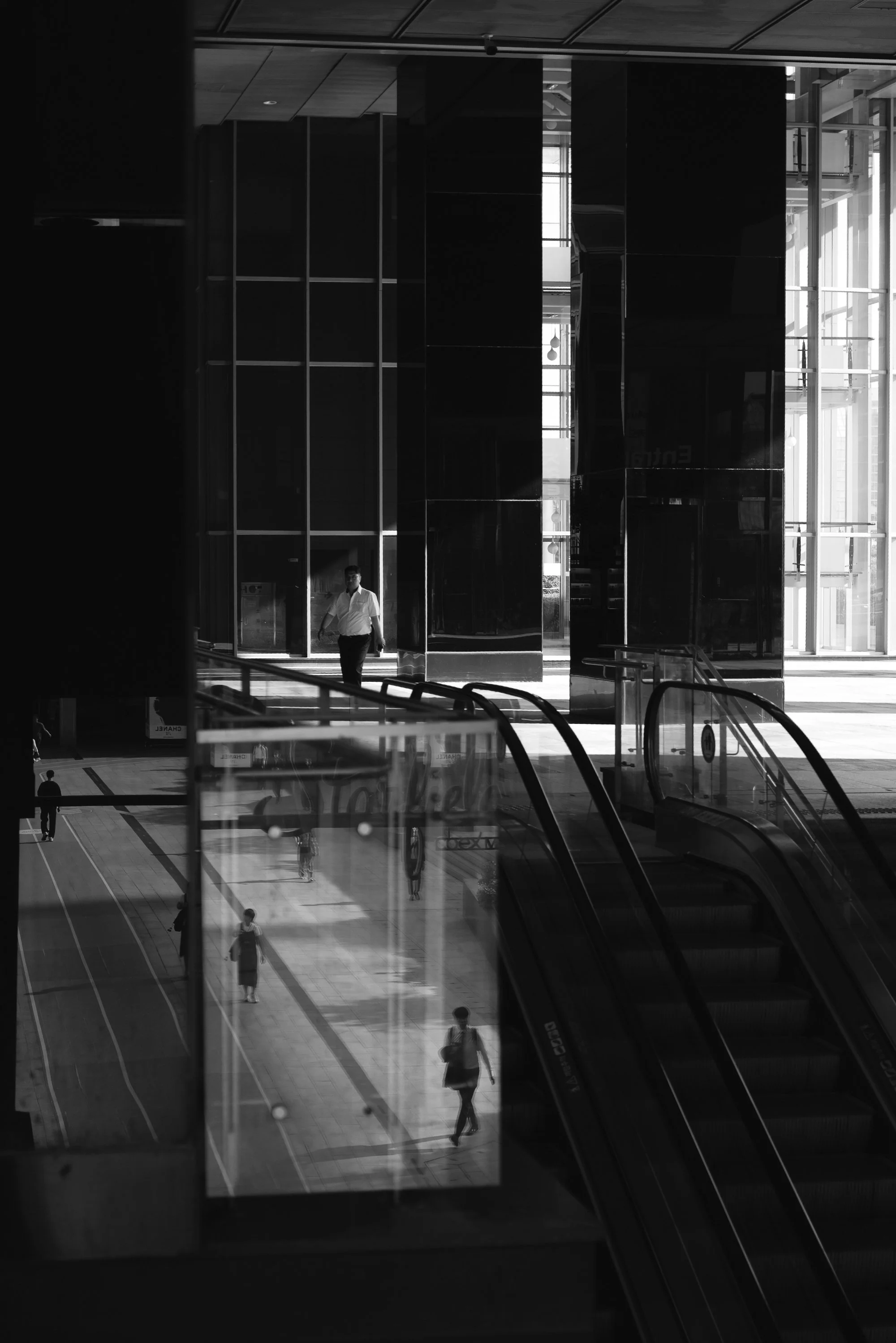 Print AD-0018. People among escalators, reflections and tall interior columns.