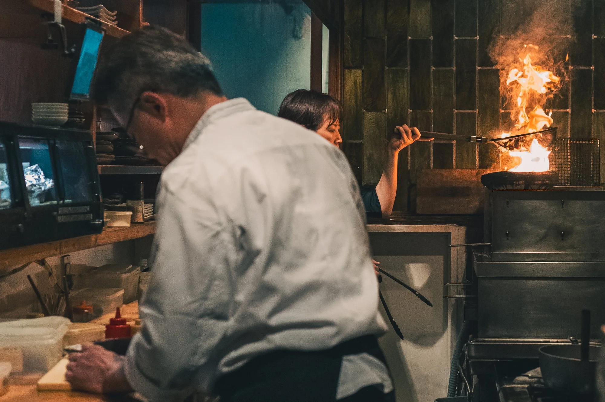 Print AD-0051. A cook preparing food while another tends a flame behind.