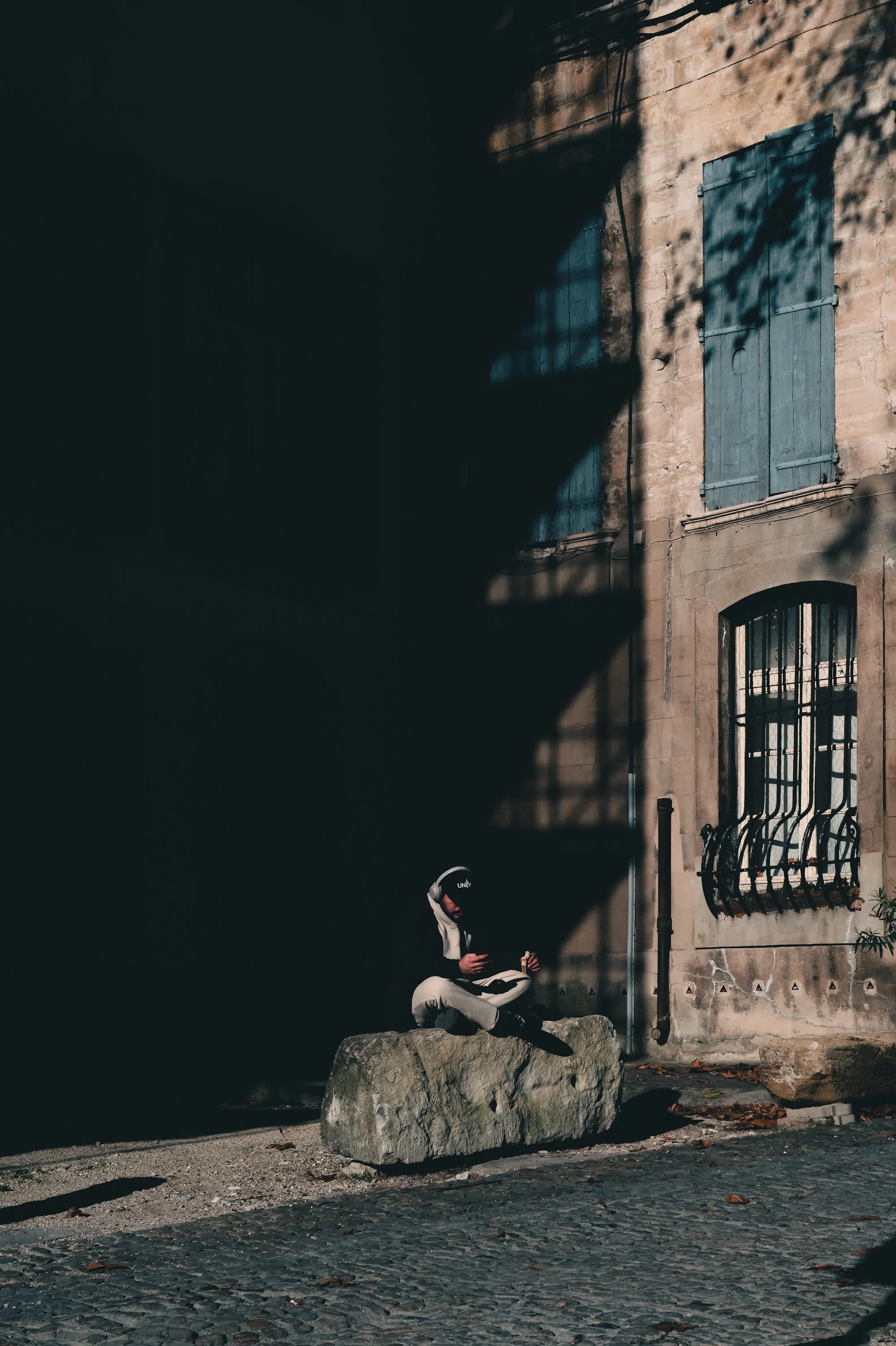 Print AD-0022. A seated person at the edge of light beside a shuttered building.