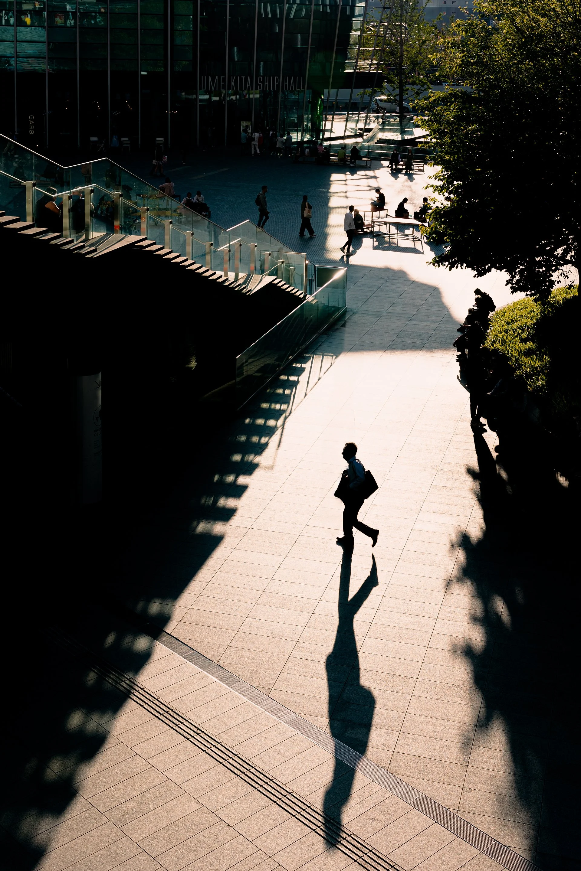 Print AD-0020. A walking figure and long shadow in a sunlit public square.