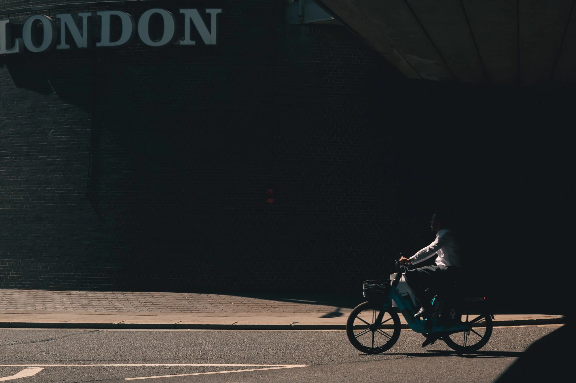 Print AD-0021. A cyclist crossing a strip of sunlight beside a dark brick wall.