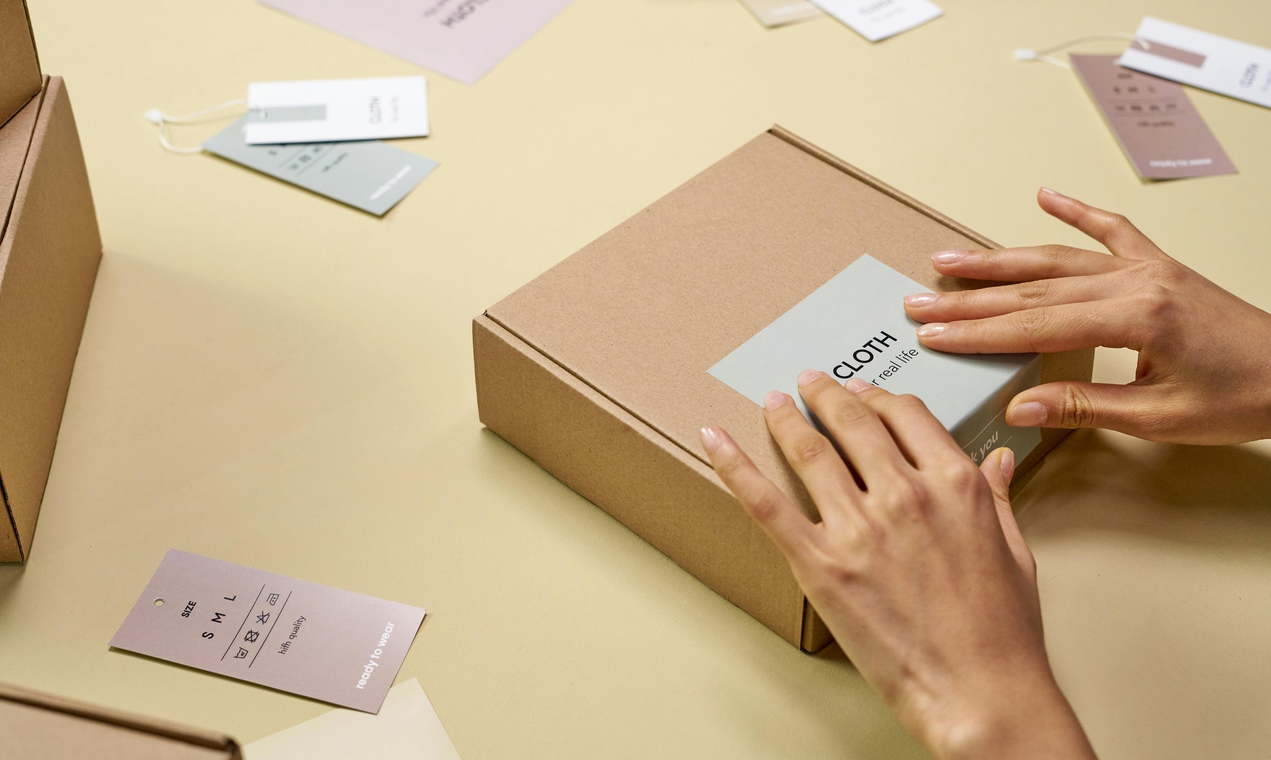 Hands placing a branded label on a cardboard product box surrounded by clothing tags on a beige surface.