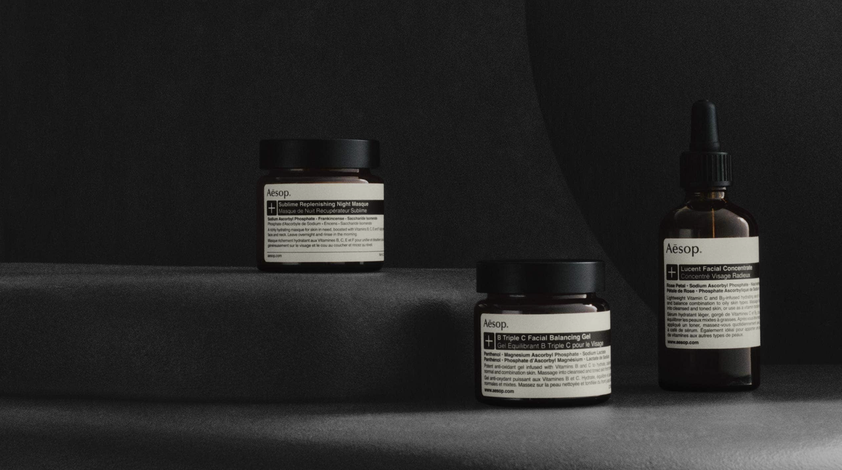 Minimalist display of three Aesop skincare products, including jars and a dropper bottle, arranged on dark fabric with soft lighting.