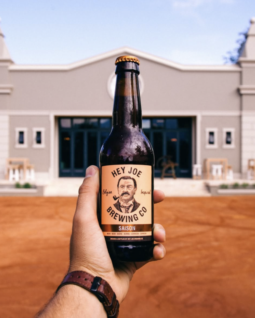 Hand holding a Hey Joe Brewing Co beer bottle with a portrait label, in front of a modern brewery building.