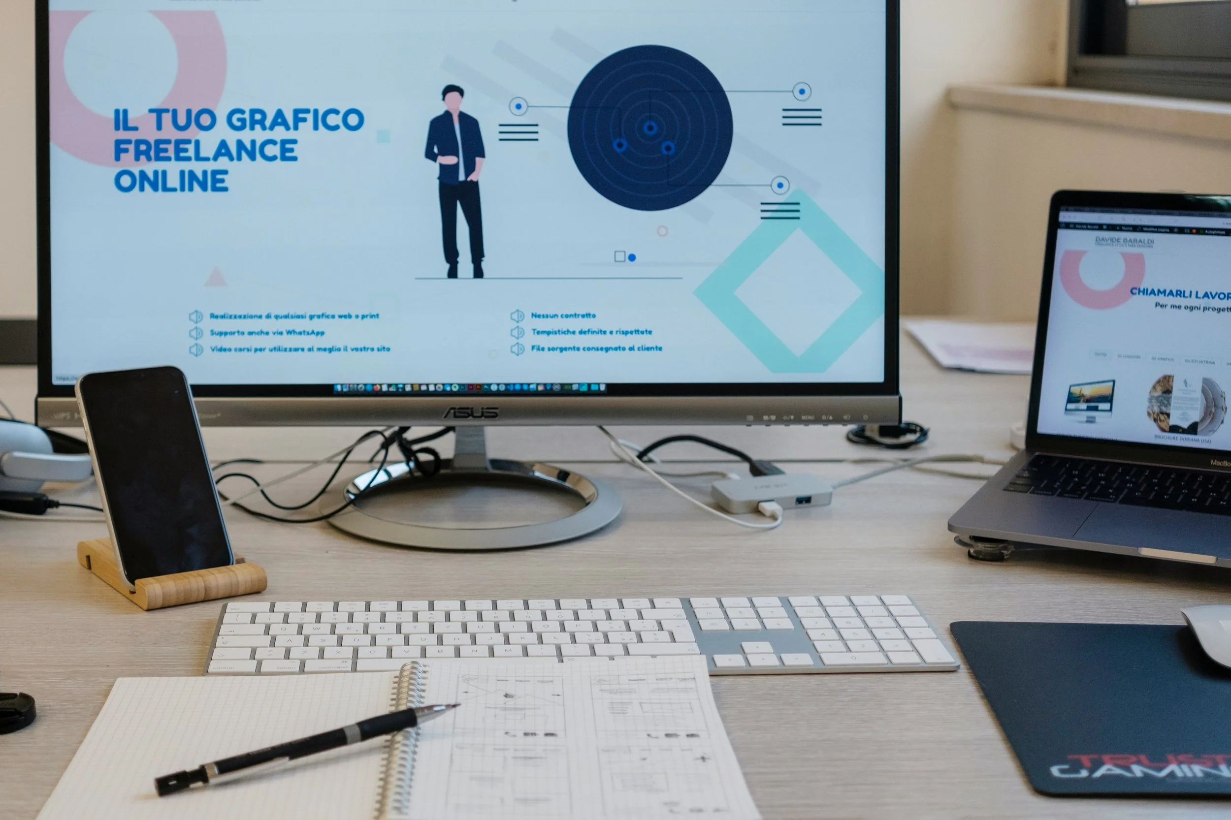 Desktop setup with a large monitor showing a freelance graphic design website in Italian, a laptop, notebook, pen, and smartphone on a wooden stand.
