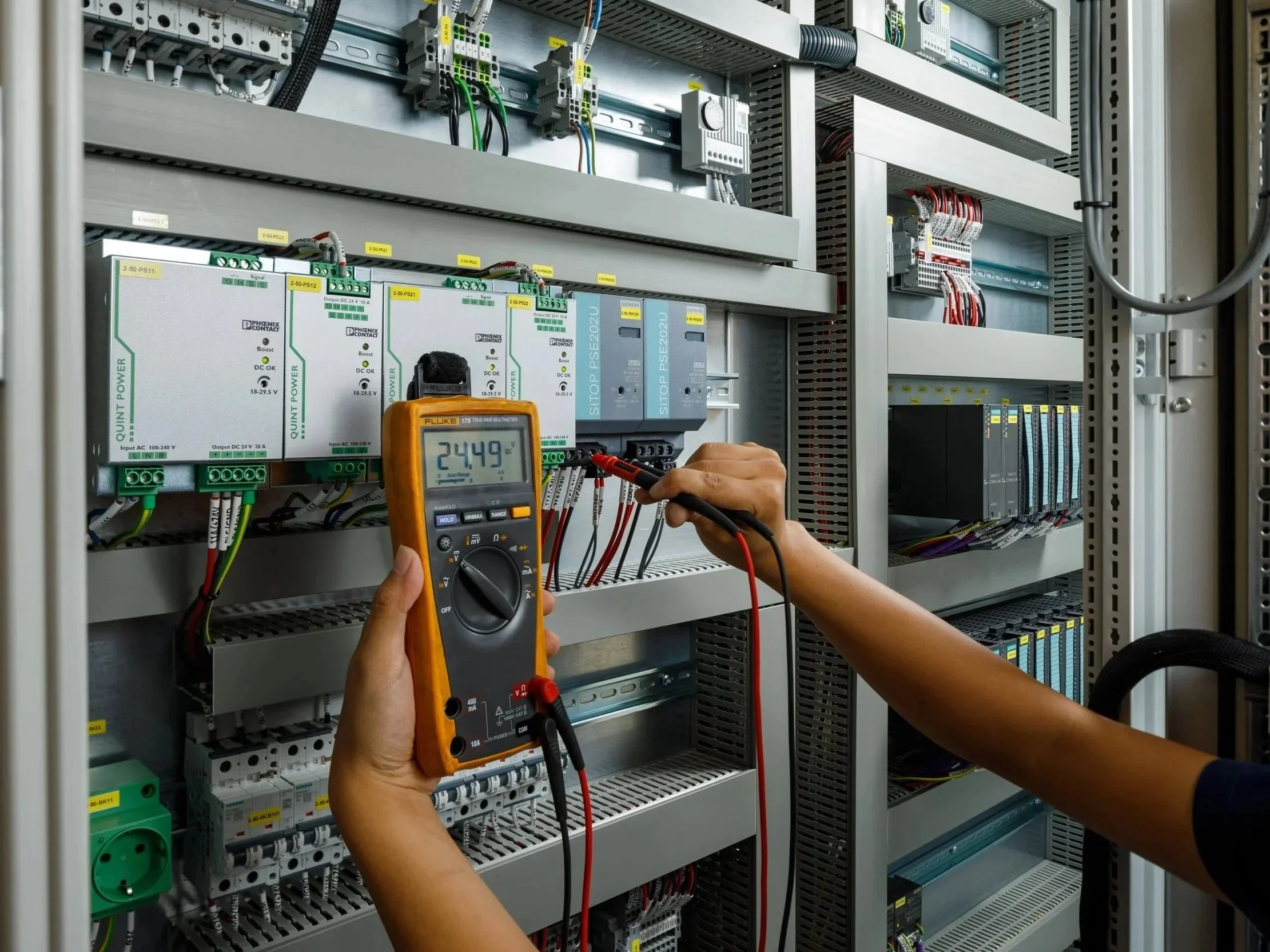 Technicians working on an electrical control panel, using a multimeter to test circuitry inside a metal enclosure filled with wires and electronic components.
