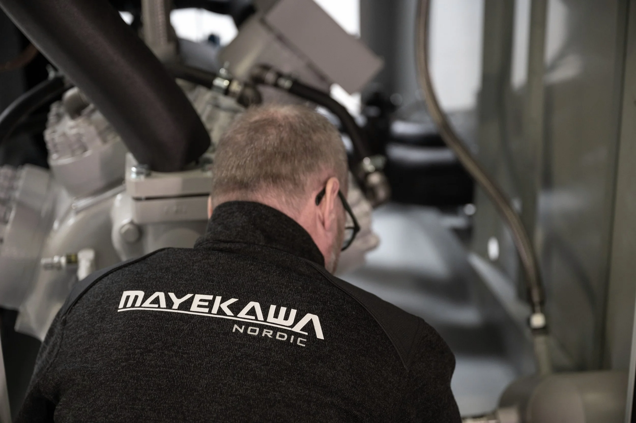 A man viewed from behind wearing a dark jacket with the text 'MAYEKAWA NORDIC' visible on the back. He is working with machinery that appears to be part of an industrial or mechanical setting, with visible pipes, wires, and metal components.