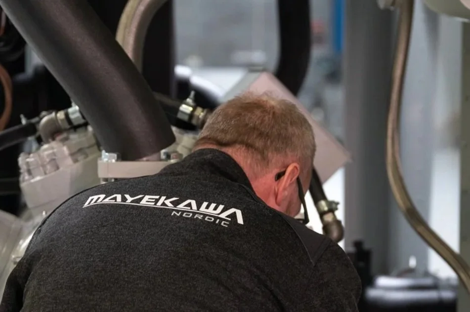 A man working on an engine in a workshop, wearing a black shirt with 'MAYEKAW A NORDIC' written on the back.