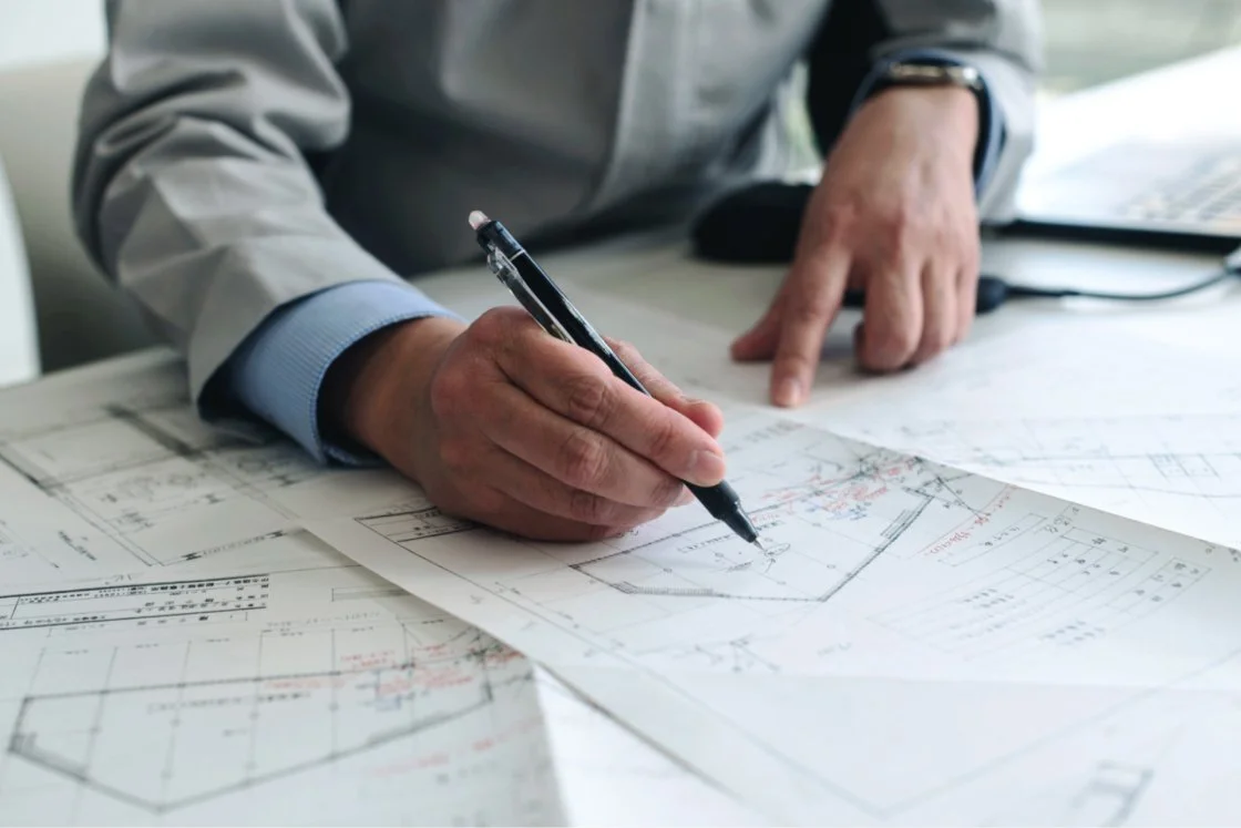 A person in a gray suit and blue shirt is drawing on architectural blueprints with a black pen, with a laptop nearby.