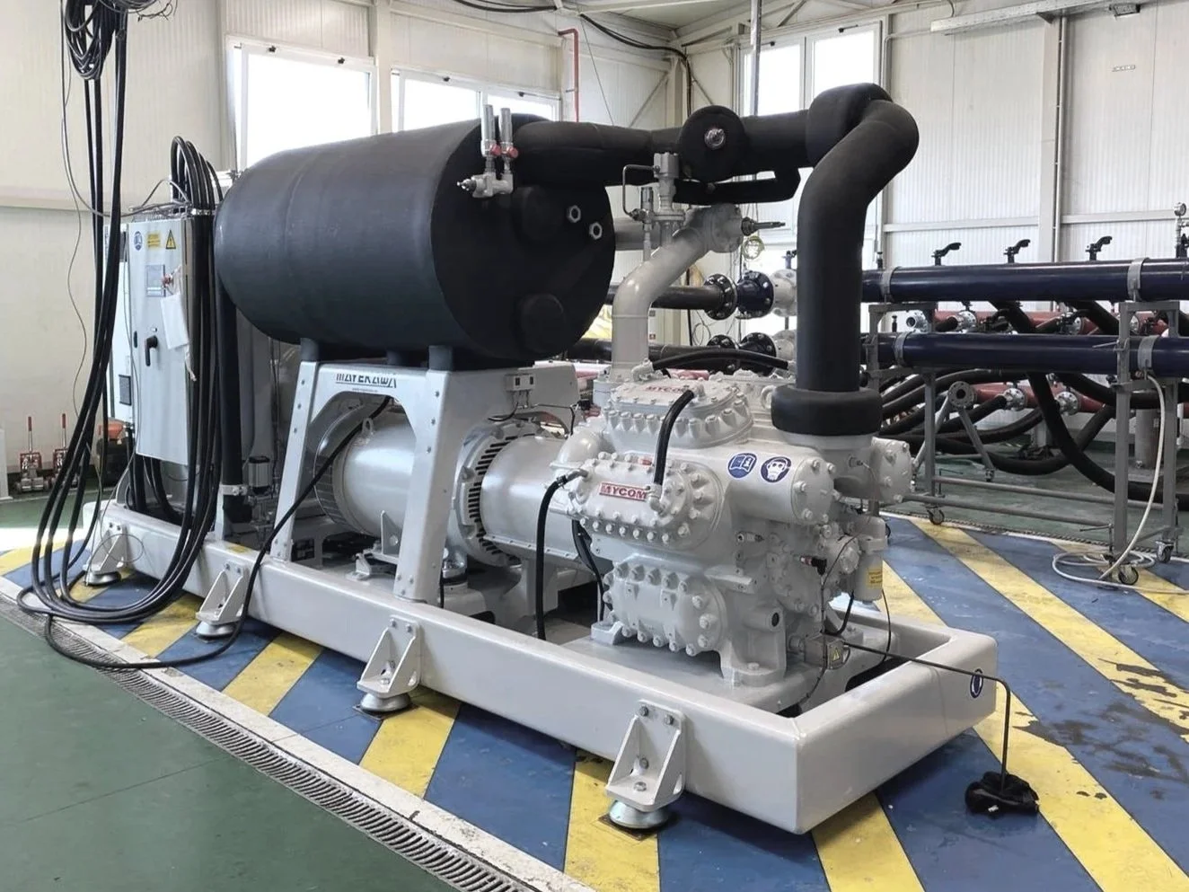 Industrial equipment with black cylindrical components, pipes, valves, and electrical cables inside a factory or plant.