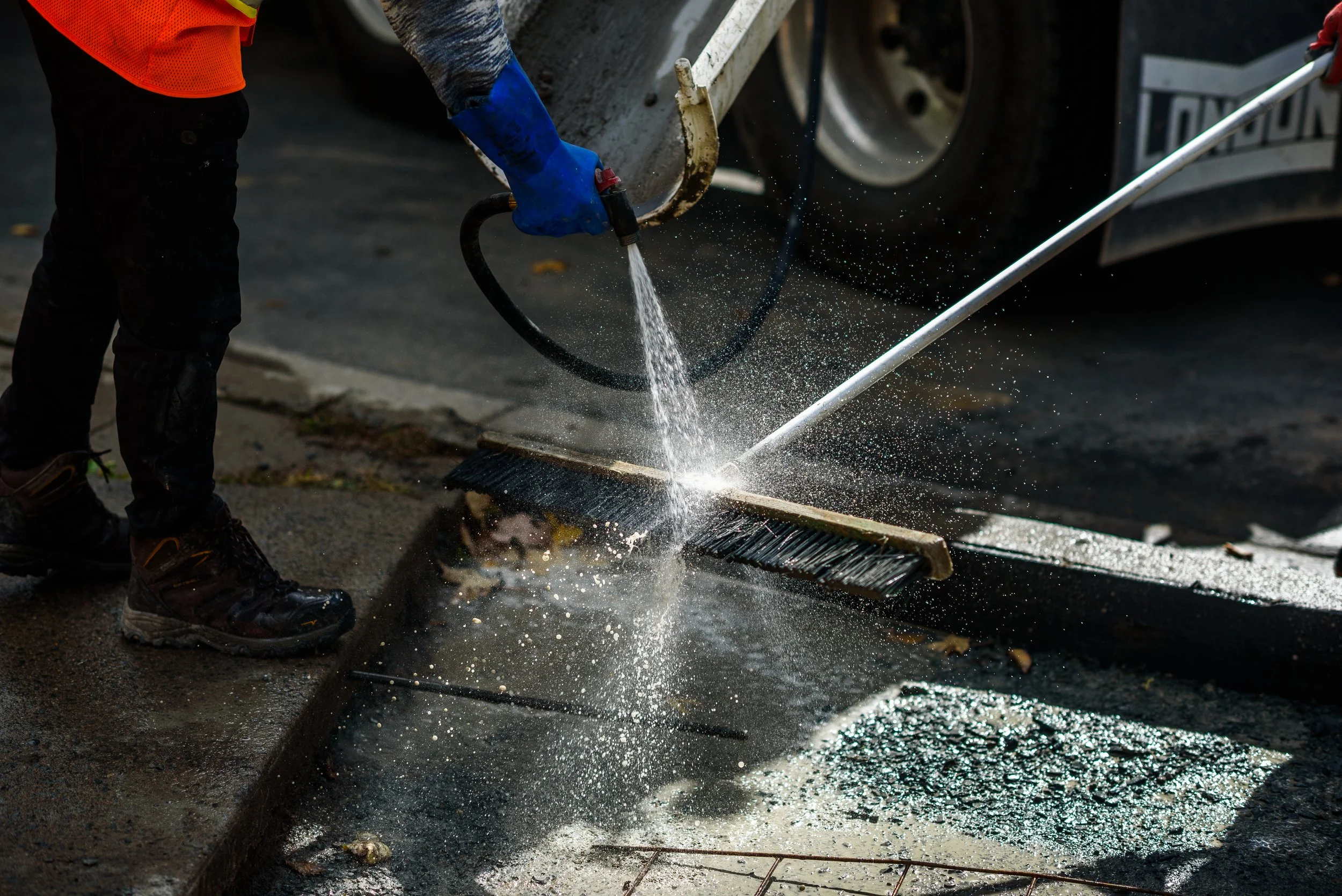 Worker using a power washer to clean a sidewalk, with broken glass scattered on the ground.