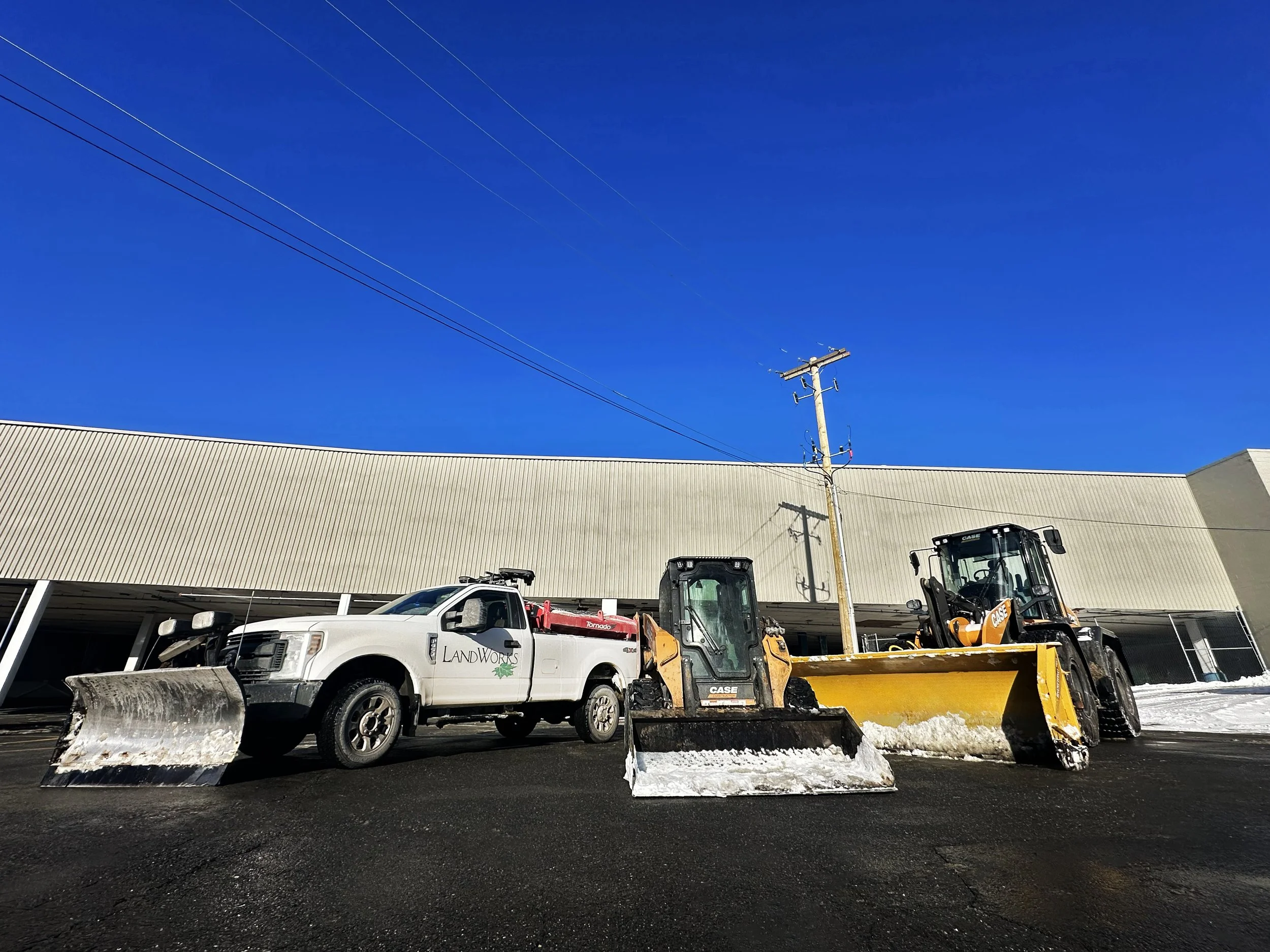 Construction vehicles, including a white pickup truck with a snow plow and two yellow snow removal machines, parked on a road in front of a beige building with a large flat roof under a blue sky.