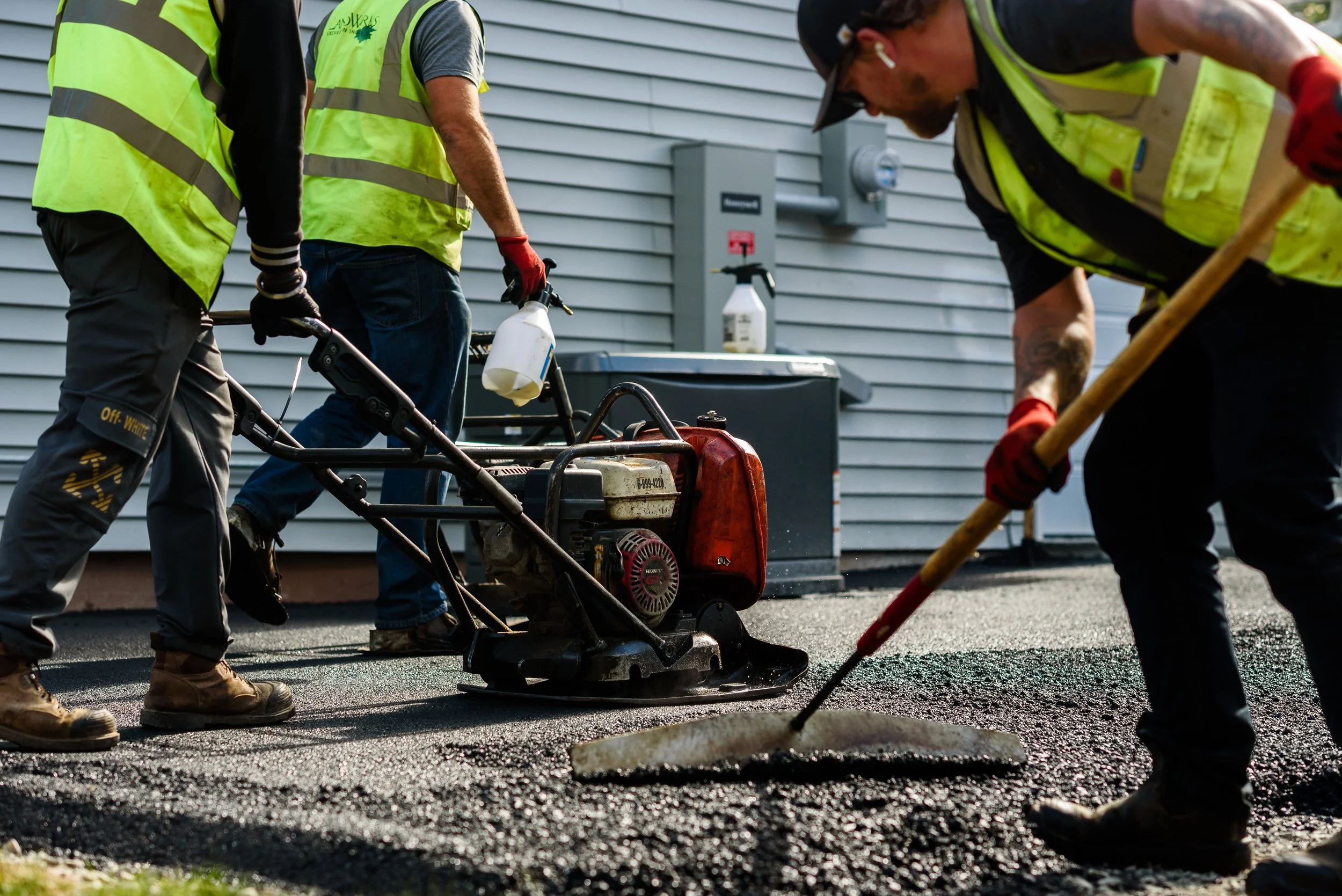 Workers in yellow safety vests and red gloves paving a road with asphalt using heavy equipment and hand tools.