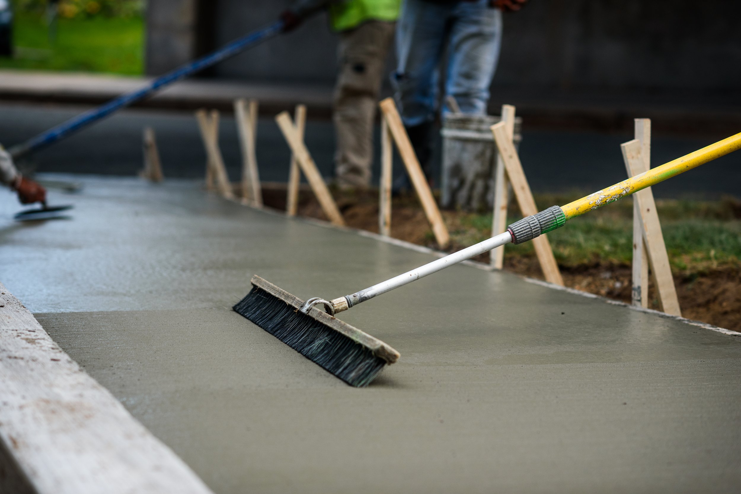 Workers smoothing wet concrete with a long-handled tool at a construction site.