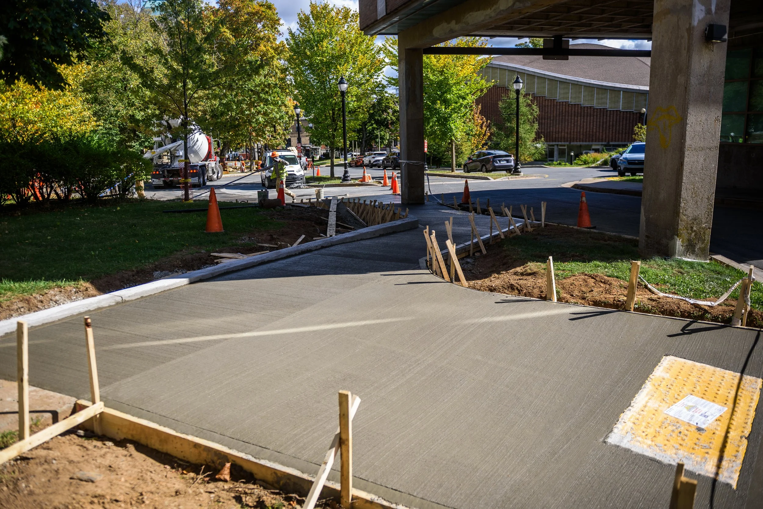 Construction workers installing a new sidewalk beneath an overpass, with orange cones, wooden stakes, and construction equipment visible.