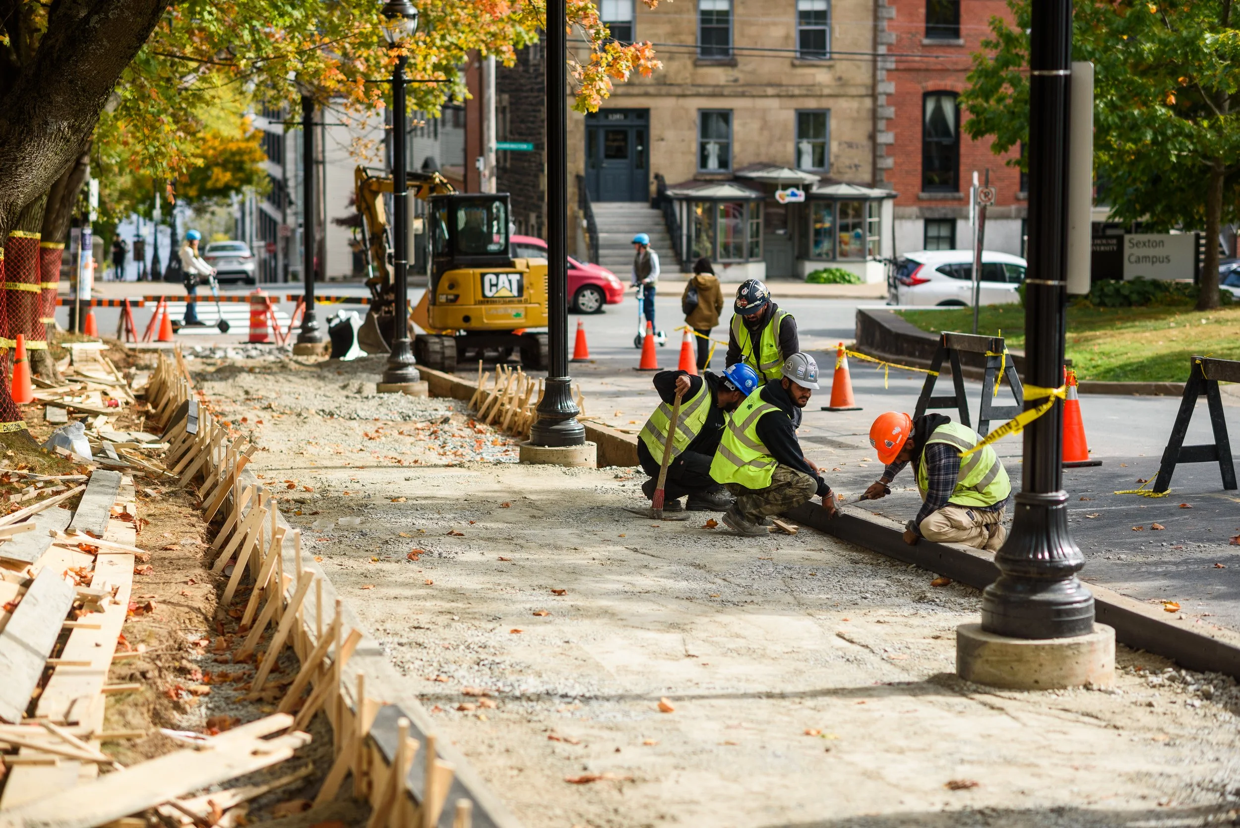Construction workers in safety vests and helmets working on sidewalk renovation, placing paving materials, with construction equipment and traffic cones nearby, in an urban area.
