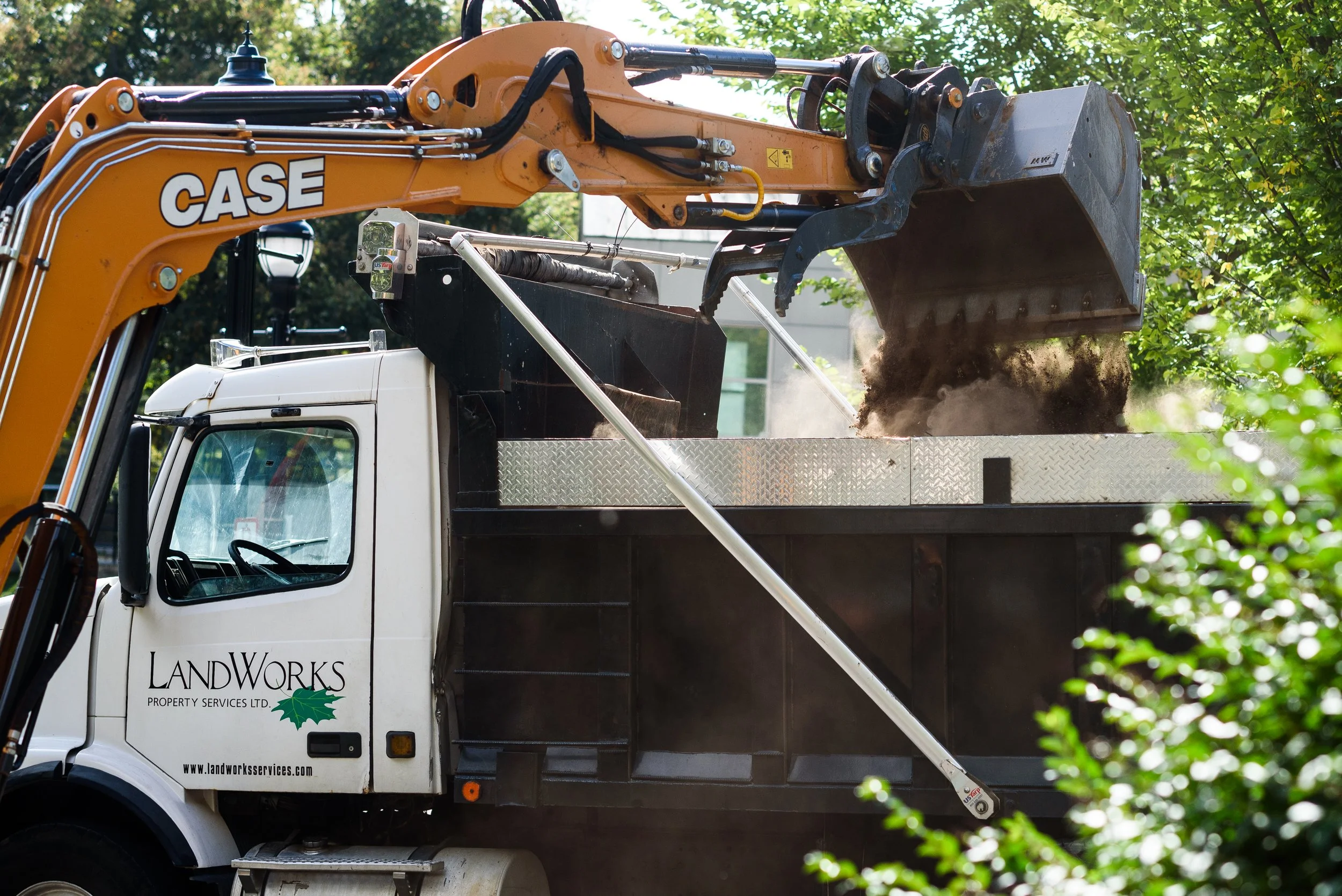 A construction truck with a hydraulic excavator arm lifting soil or dirt into the truck bed. The truck has the logo 'LandWorks Property Services Ltd.' on the door, and the excavator arm has the brand name 'CASE'.
