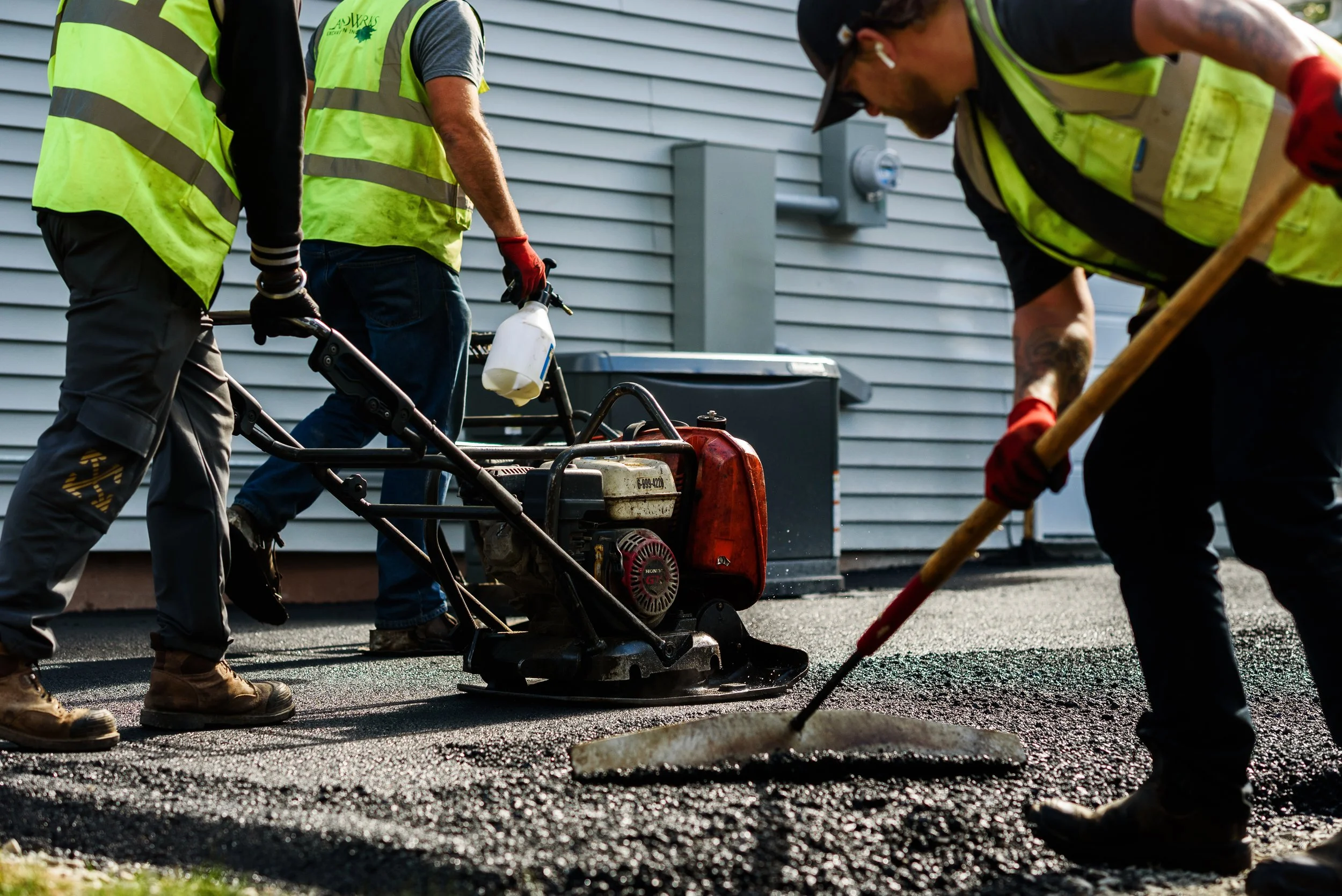 Three workers in safety vests and gloves paving a driveway with a road roller and a rake.