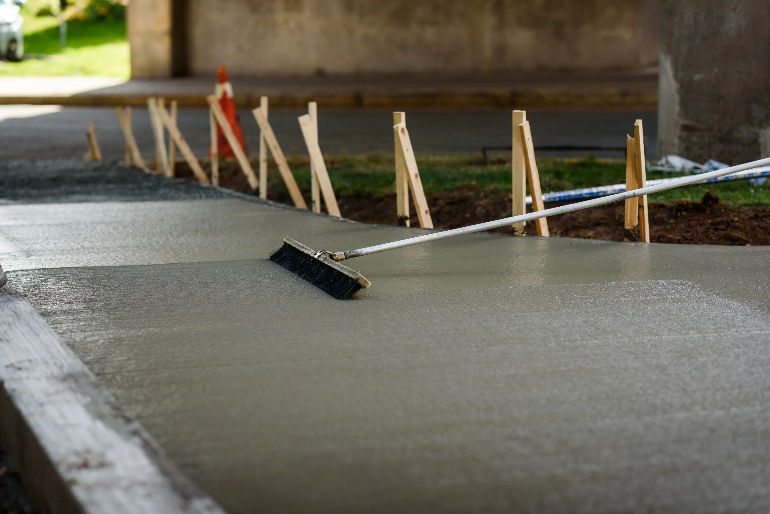 Concrete being poured and leveled on a construction site with wooden stakes and a broom resting on the wet surface.