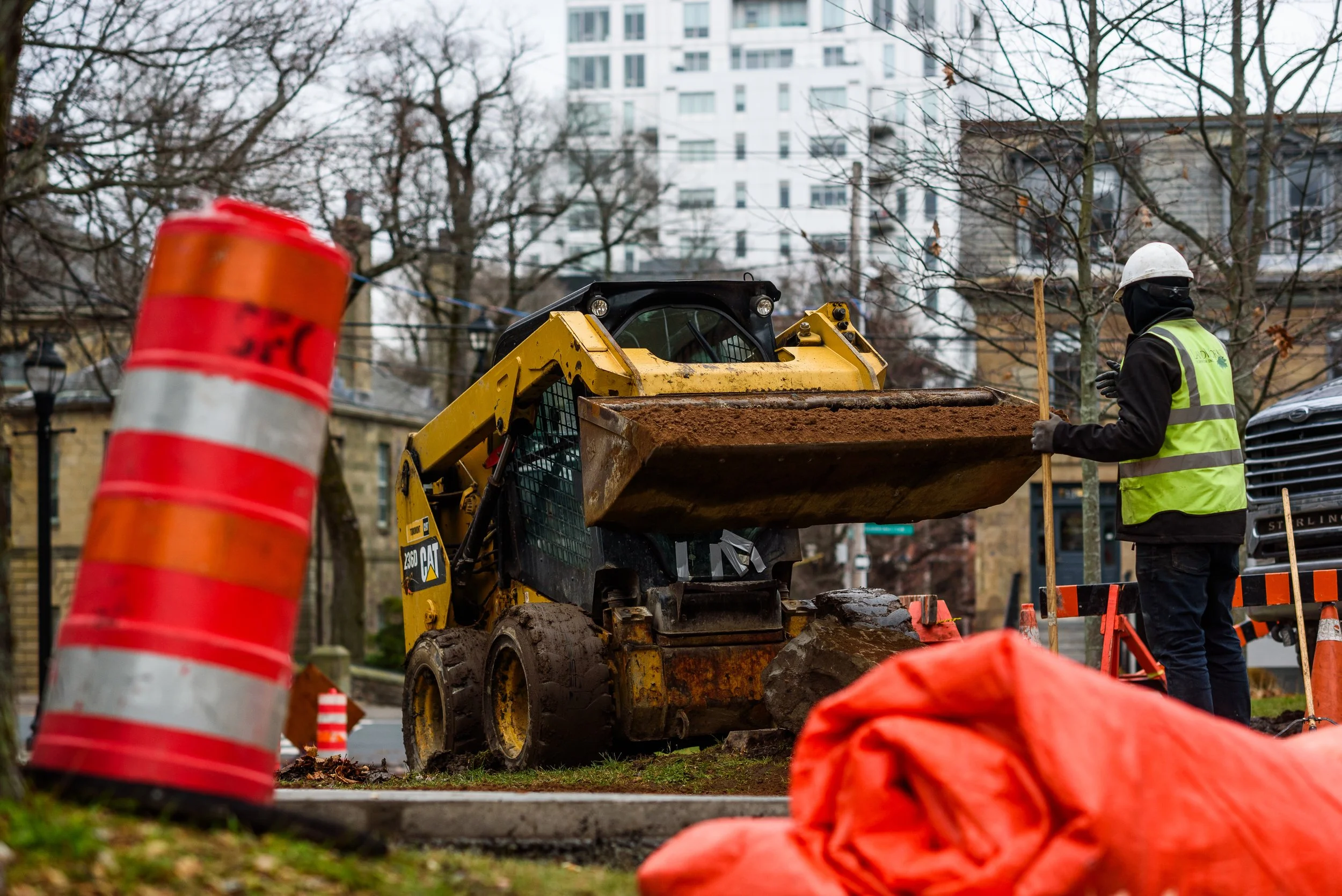 A construction worker with a white helmet and yellow safety vest standing near a small yellow Caterpillar skid-steer loader. The worker is holding a wooden stick, and the loader has dirt in its bucket. Orange safety cones and a large orange bag are i