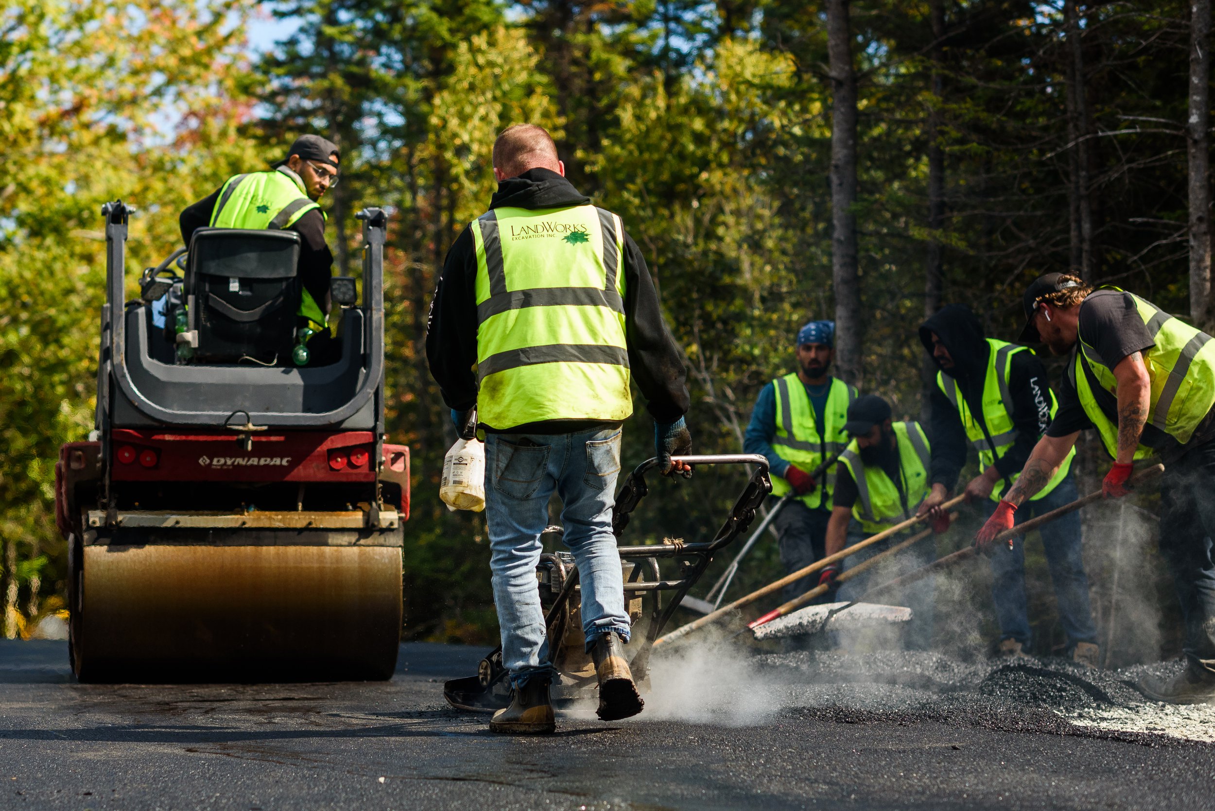 Workers in high-visibility safety vests paving a road with asphalt in a wooded area.
