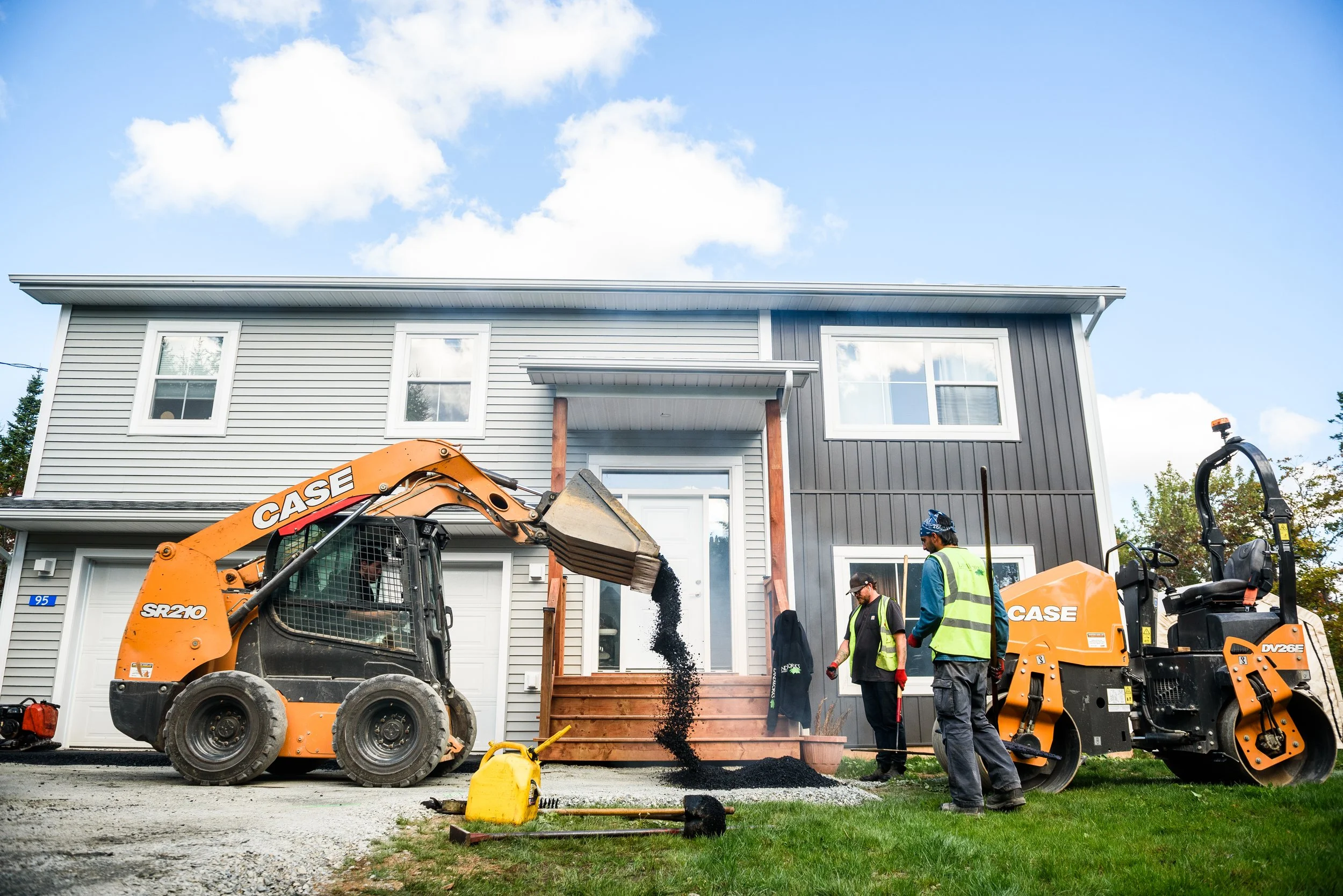 Construction workers operating machinery and working on the porch of a new two-story house with grey and black siding and a front door with a small ramp.