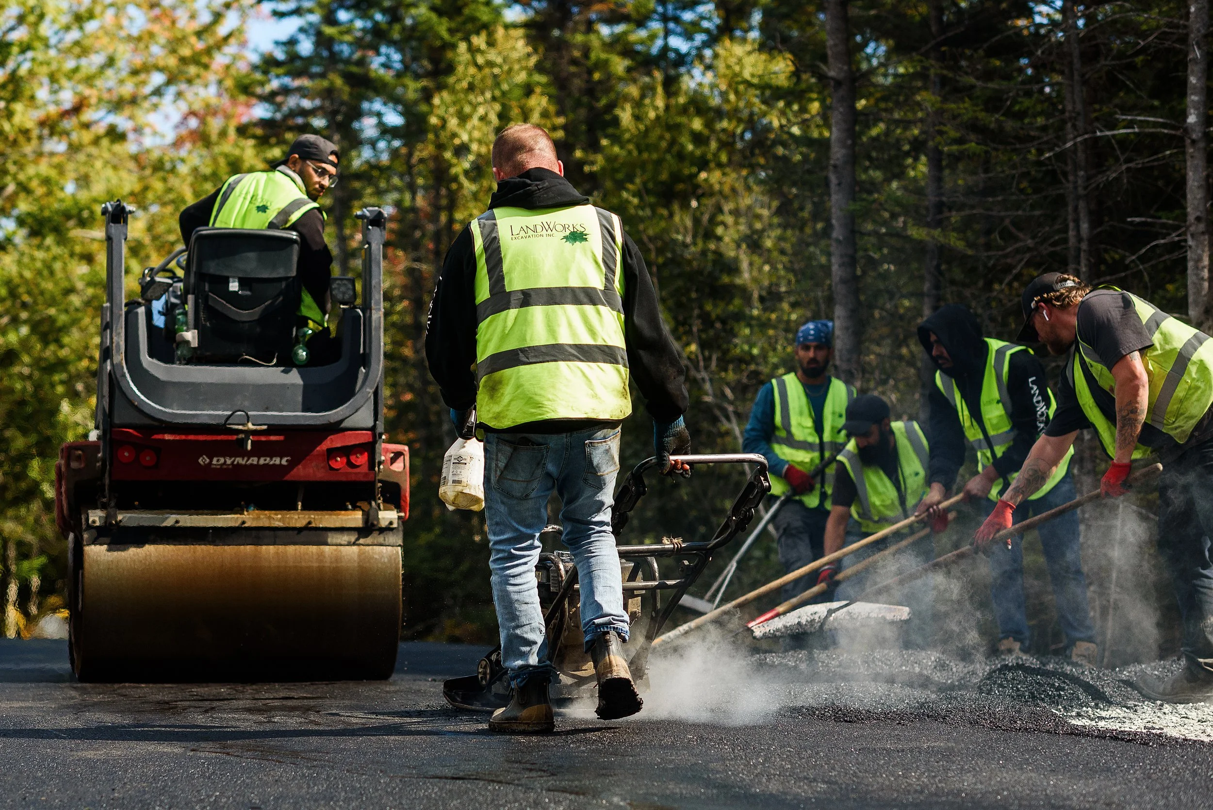 Construction workers paving a road in a forested area, wearing safety vests.