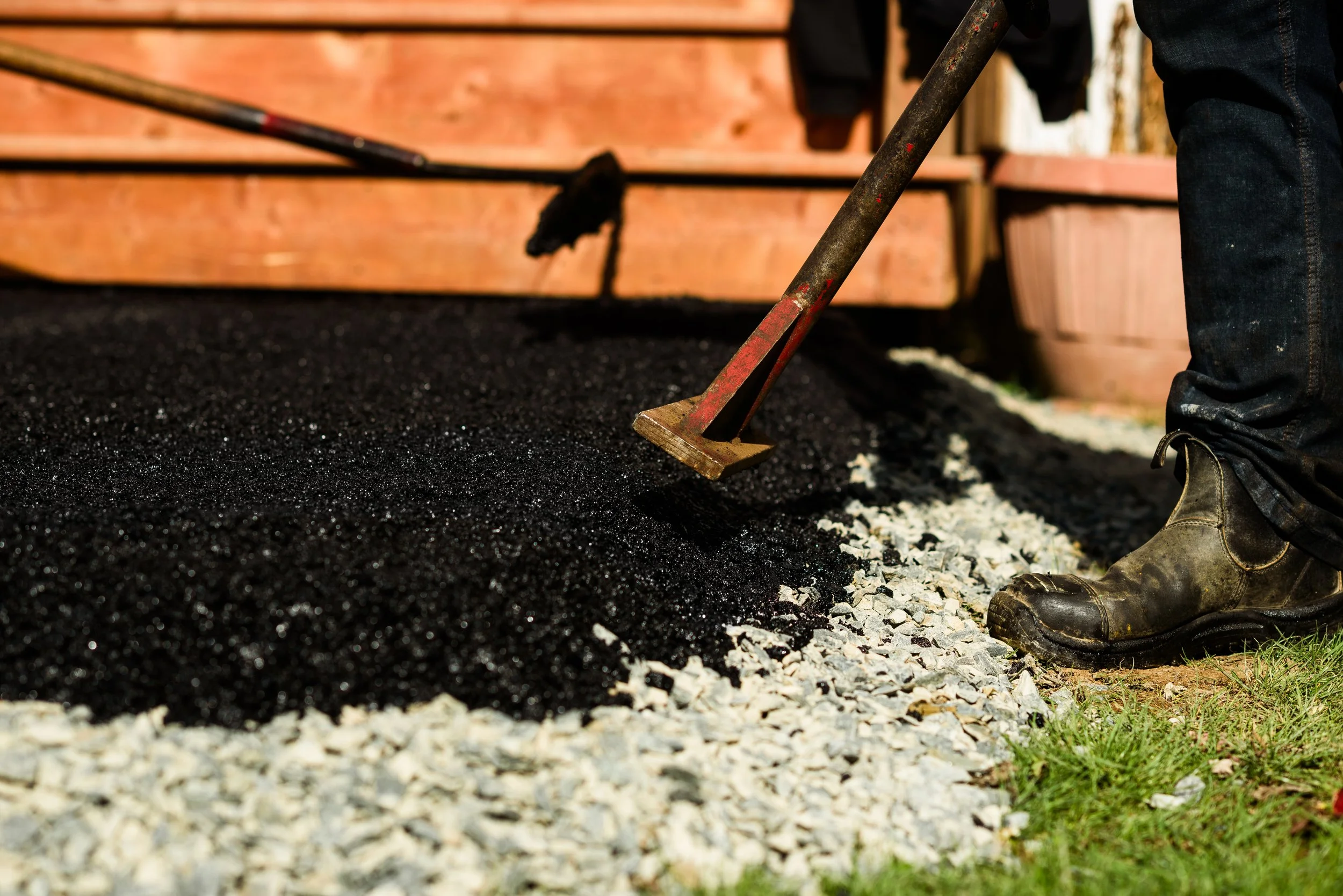 A person wearing work boots and blue jeans raking black gravel alongside white gravel outside a wooden structure.