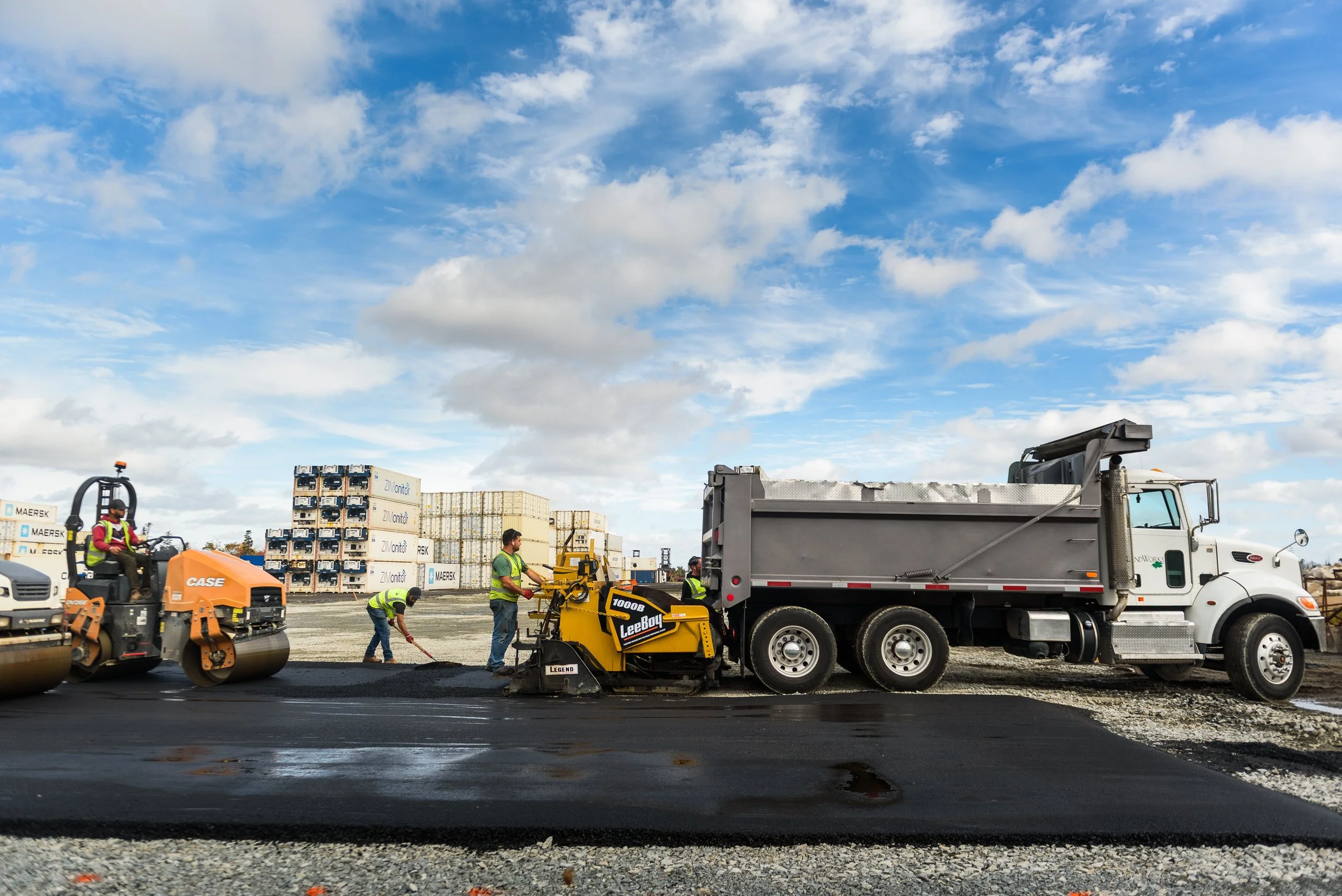 Workers paving a road with asphalt, with construction equipment and trucks in the background under a blue sky with clouds.