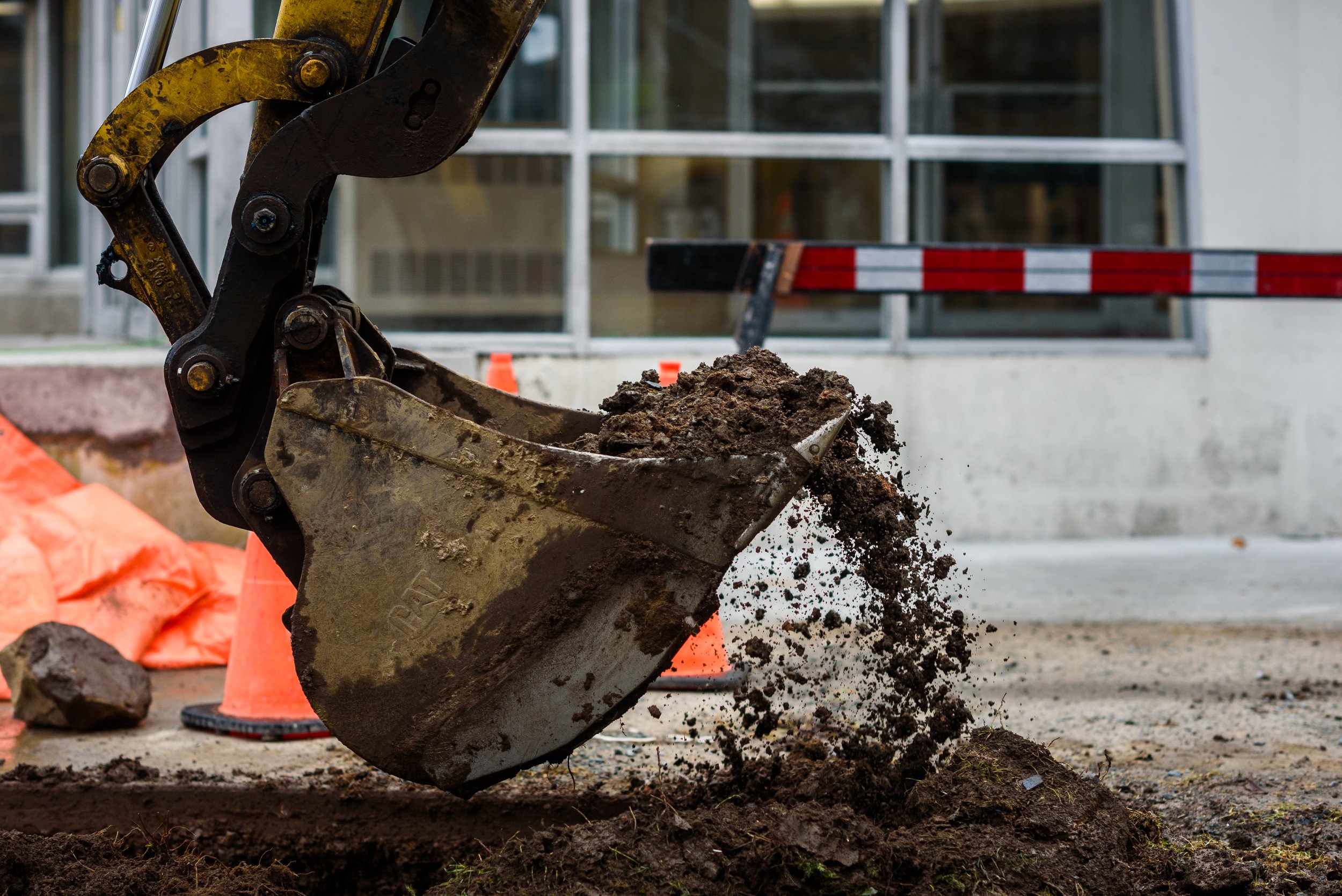 An excavator scoop lifting dirt at a construction site with orange cones and safety barriers in the background.