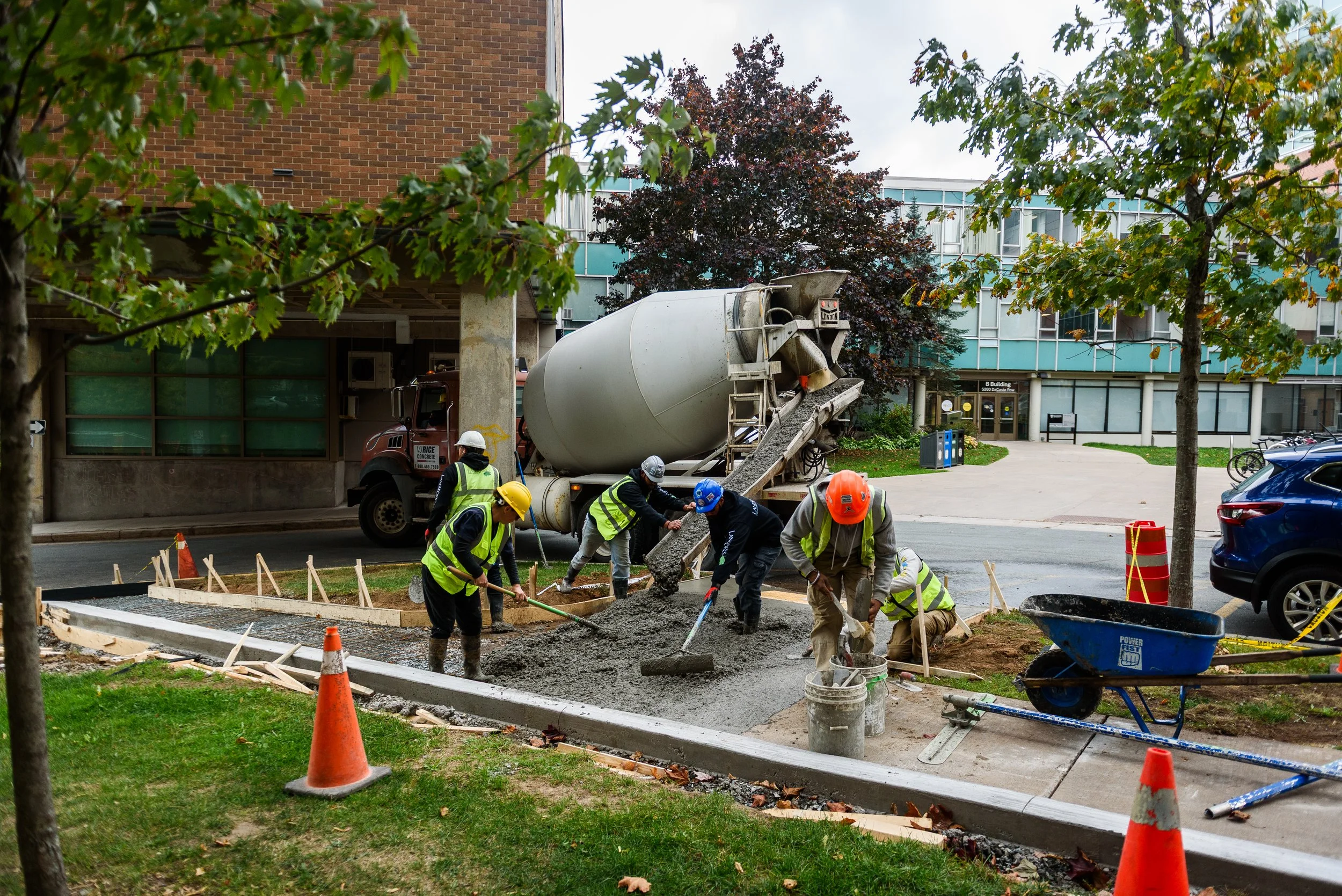 Construction workers pouring concrete for a sidewalk or pathway in an urban area, with a cement mixer truck nearby and trees around.