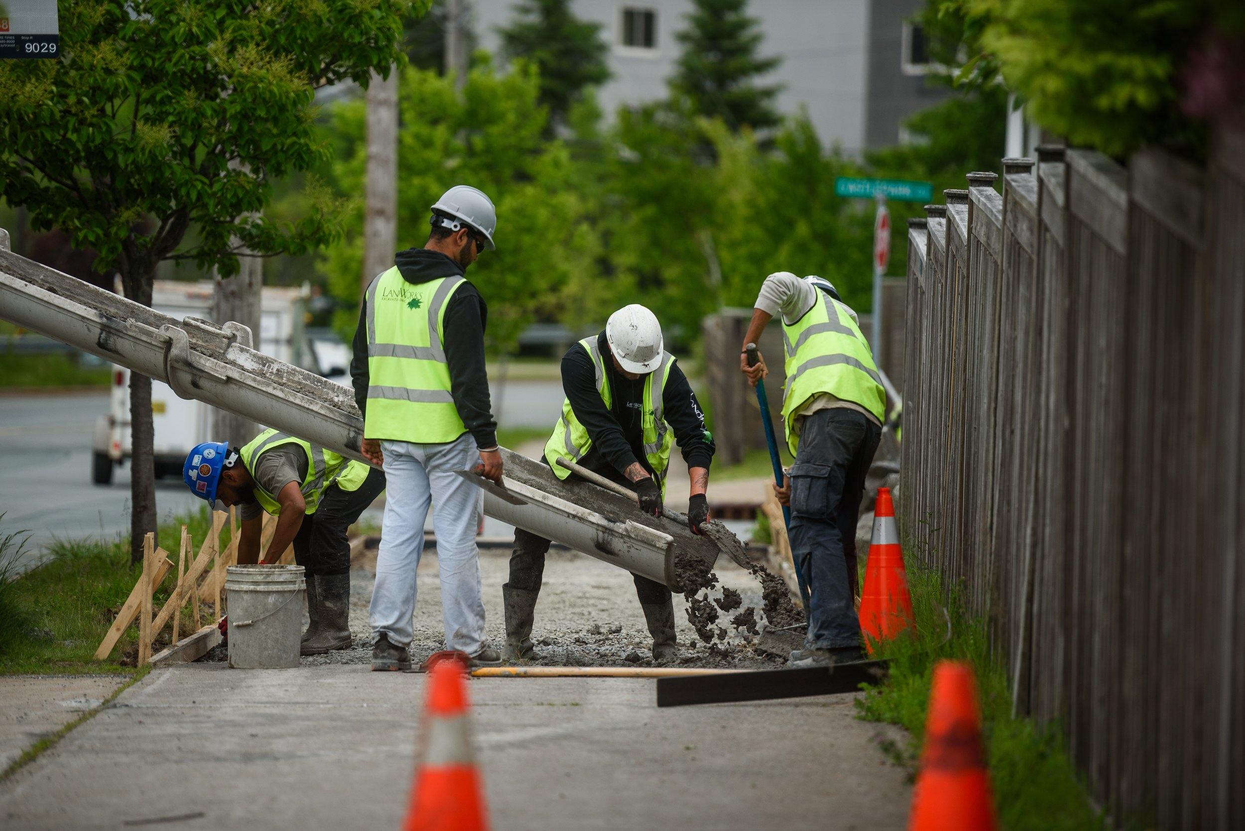 Construction workers in safety vests and helmets pouring and spreading concrete on a sidewalk, with orange safety cones and a wooden fence nearby.