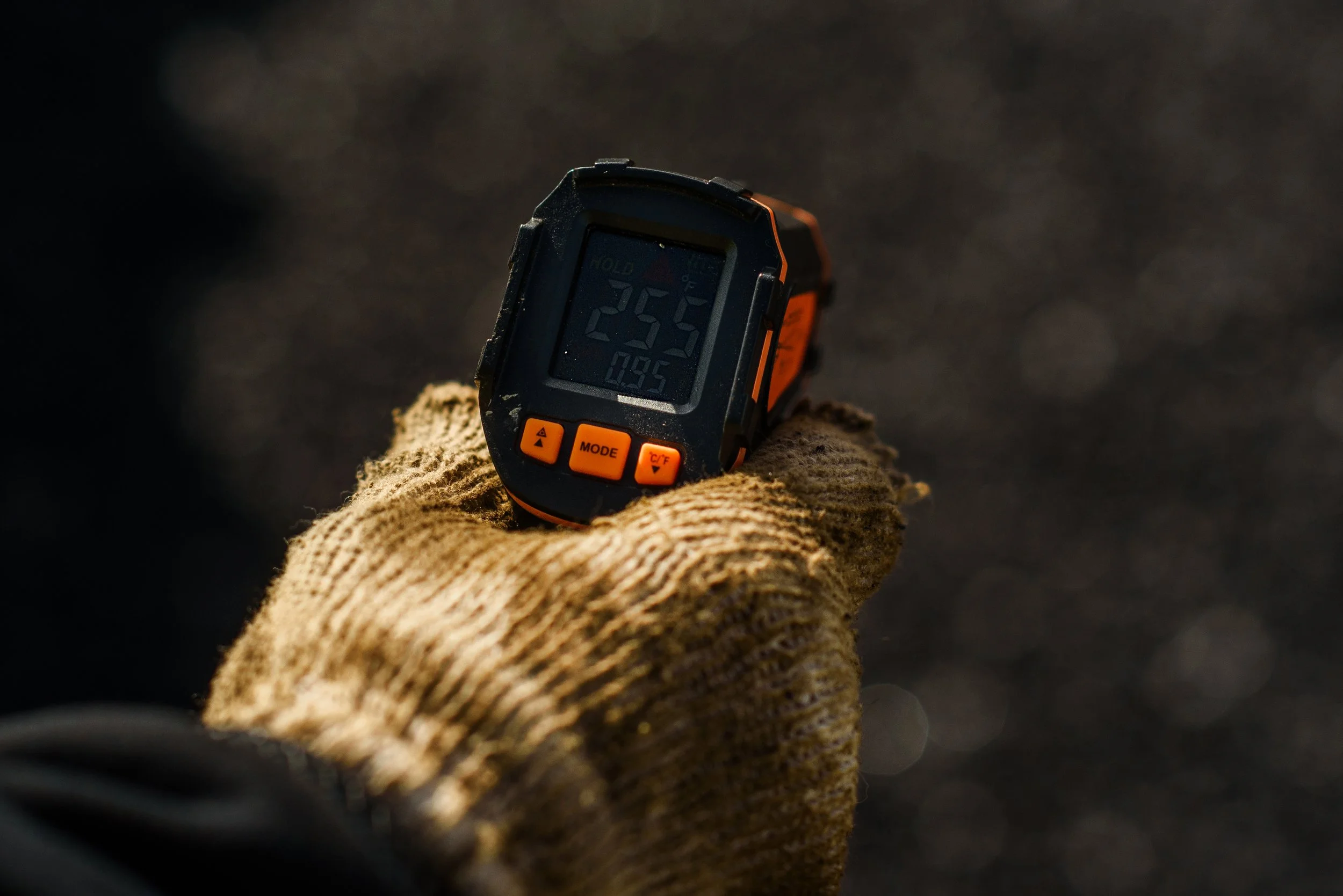 Close-up of a person's hand wearing a brown work glove, holding a digital thermometer displaying 25.5°C, against a blurred outdoor background.