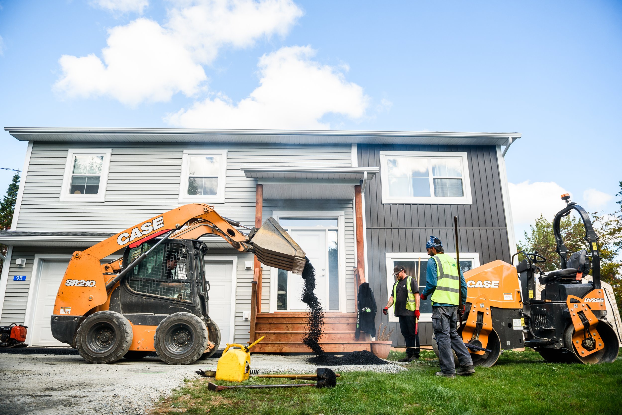 Construction workers using a skid steer loader and a roller compactor at a house renovation site, with new wooden stairs being built in front of the house.