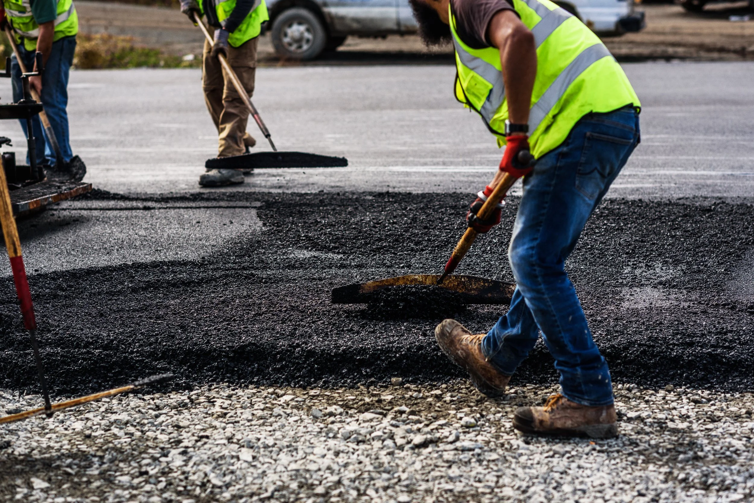 Workers in yellow safety vests and jeans resurfacing a road by spreading hot asphalt.