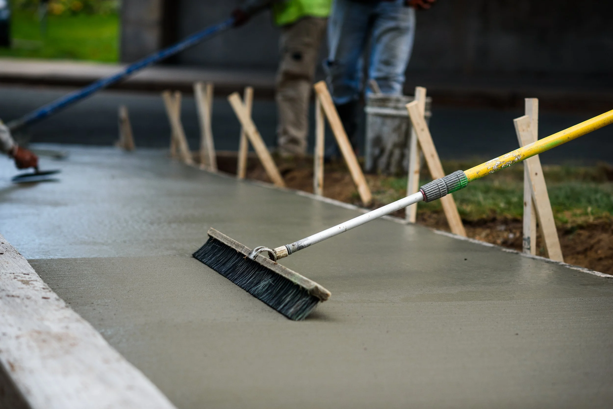 Construction workers pouring and smoothing wet concrete on a sidewalk or foundation, with wooden forms and a broom handle nearby.