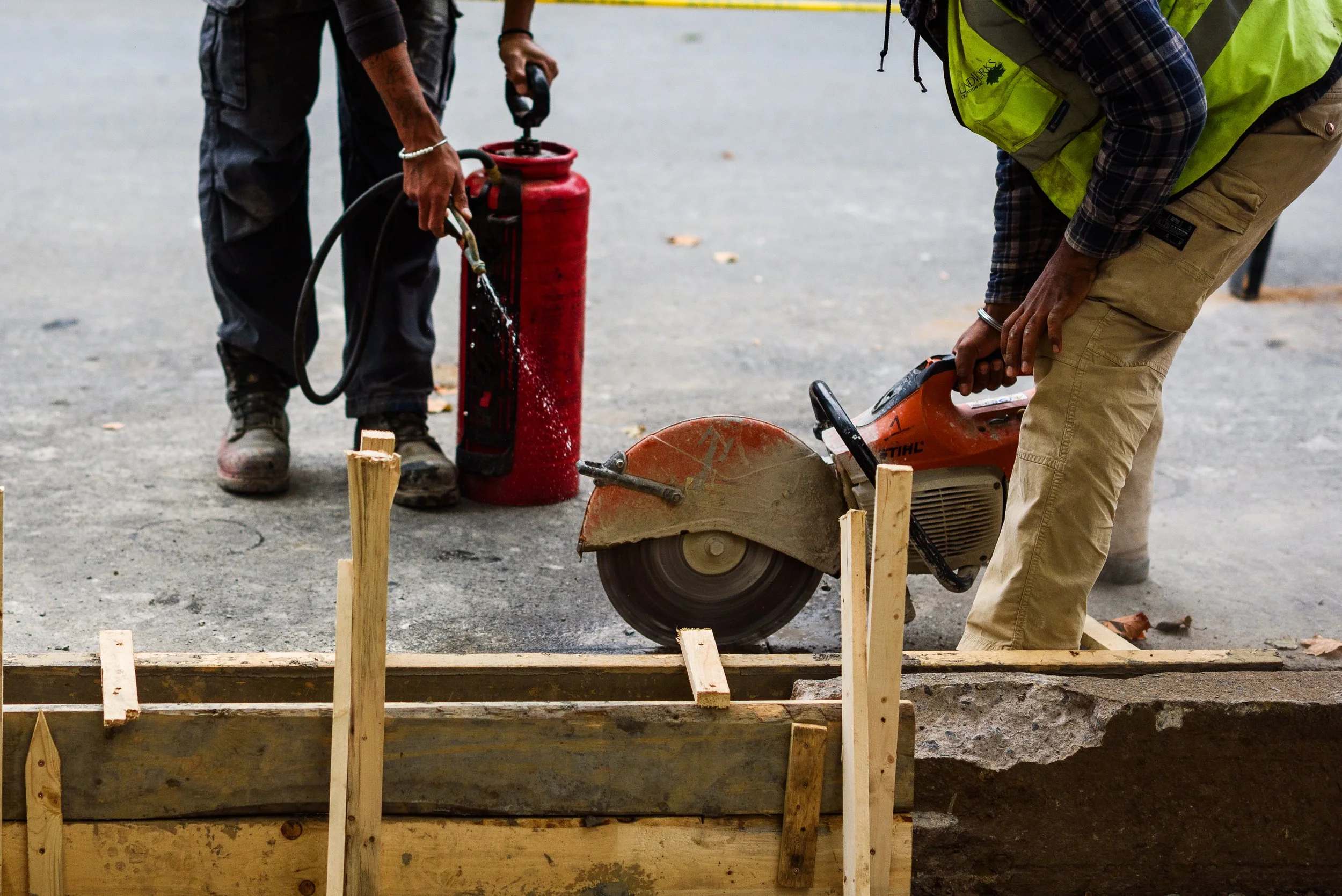 Two construction workers are using a concrete saw and a fire extinguisher during construction work on a sidewalk.