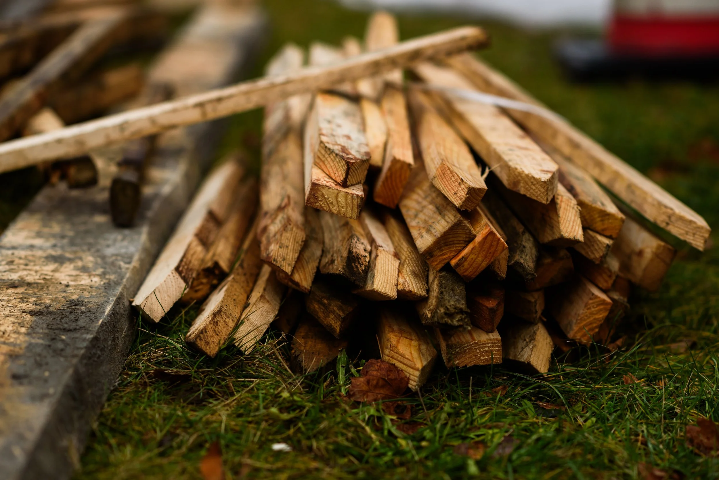 A pile of chopped firewood on the grass with some extra wood pieces nearby.