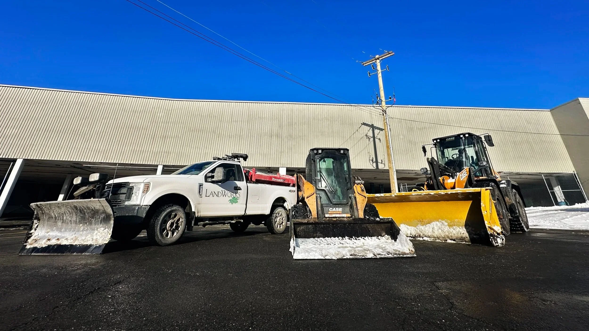 Construction vehicles including a pickup truck with a snowplow and two bulldozers parked on a freshly paved asphalt surface in front of a large industrial building with a beige metal exterior. Clear blue sky overhead.