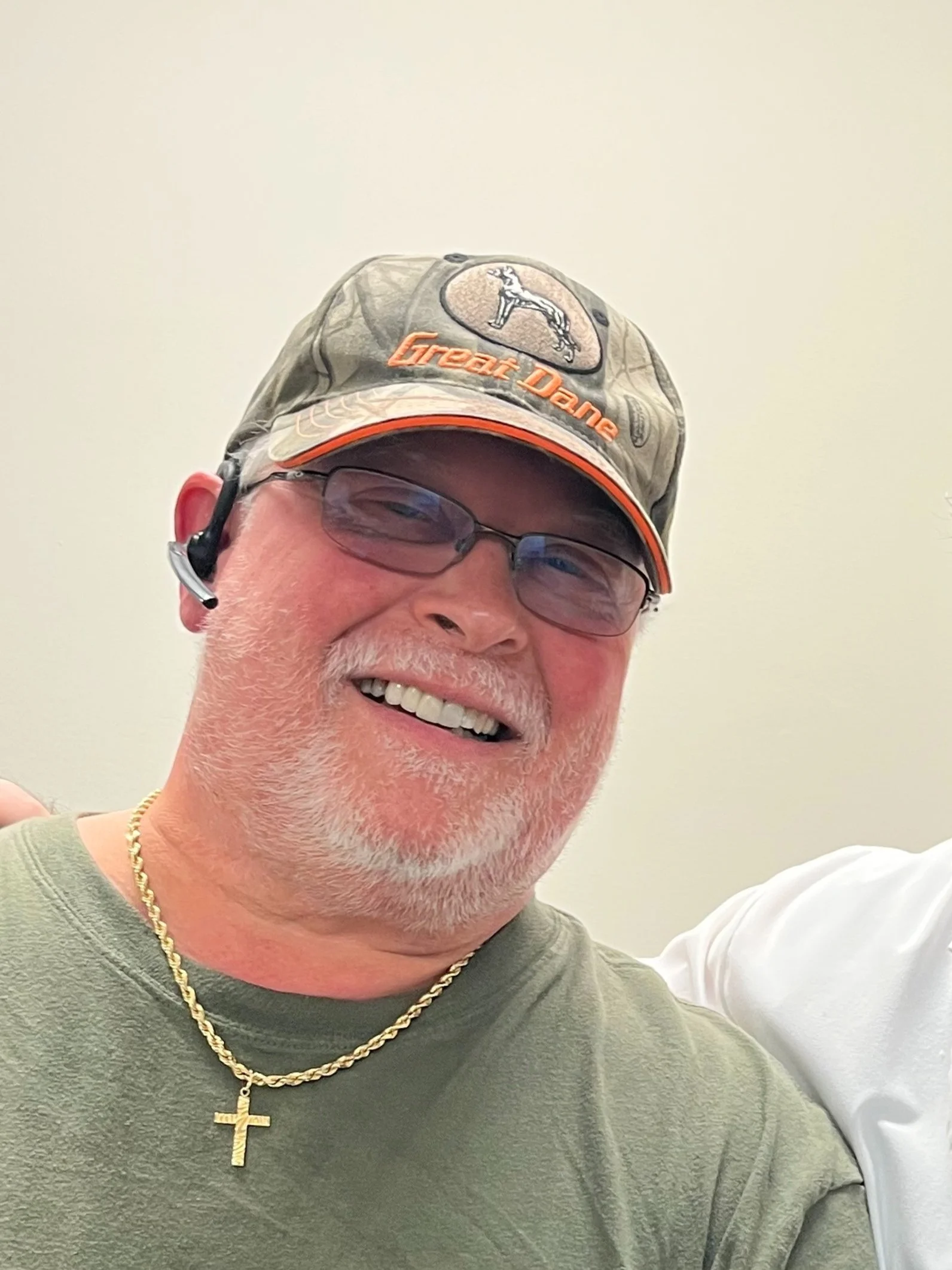 Close-up of a man smiling, wearing glasses, a camouflage baseball cap with an eagle logo and the words "GreetDane", a gold chain with a cross pendant, and a green shirt.